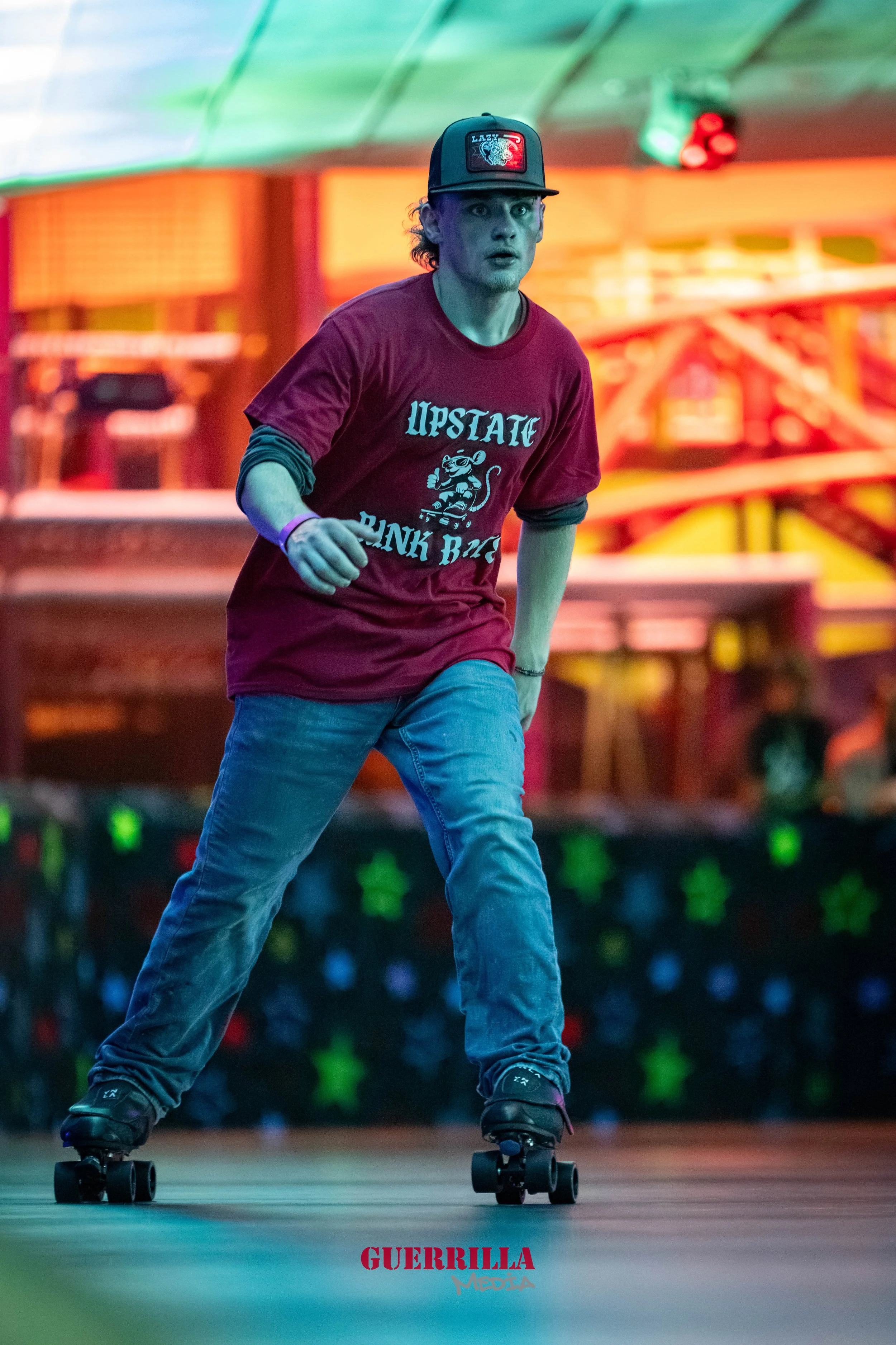 A young man riding a skateboard indoors at an amusement park or roller skating rink, wearing a maroon t-shirt with 'LIPSTATE RINK RIDE' printed on it, a black baseball cap, gray jeans, and black roller skates, with colorful lighting in the background