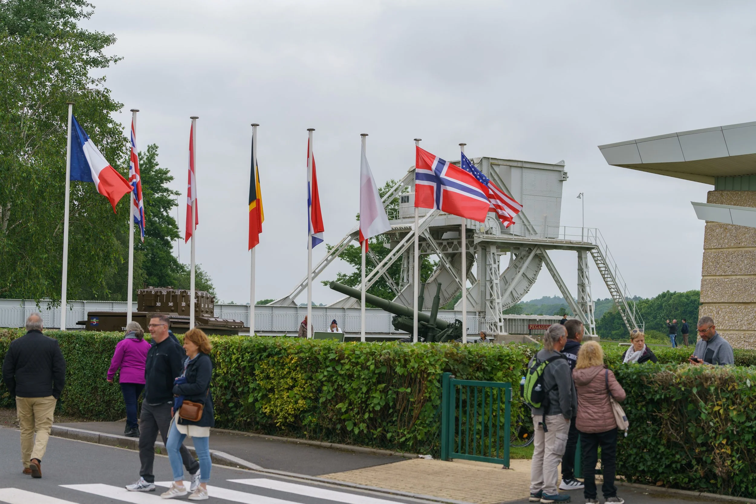 People walking past a row of flags next to a military museum with historical cannons and a large military truck, with a cloudy sky overhead.
