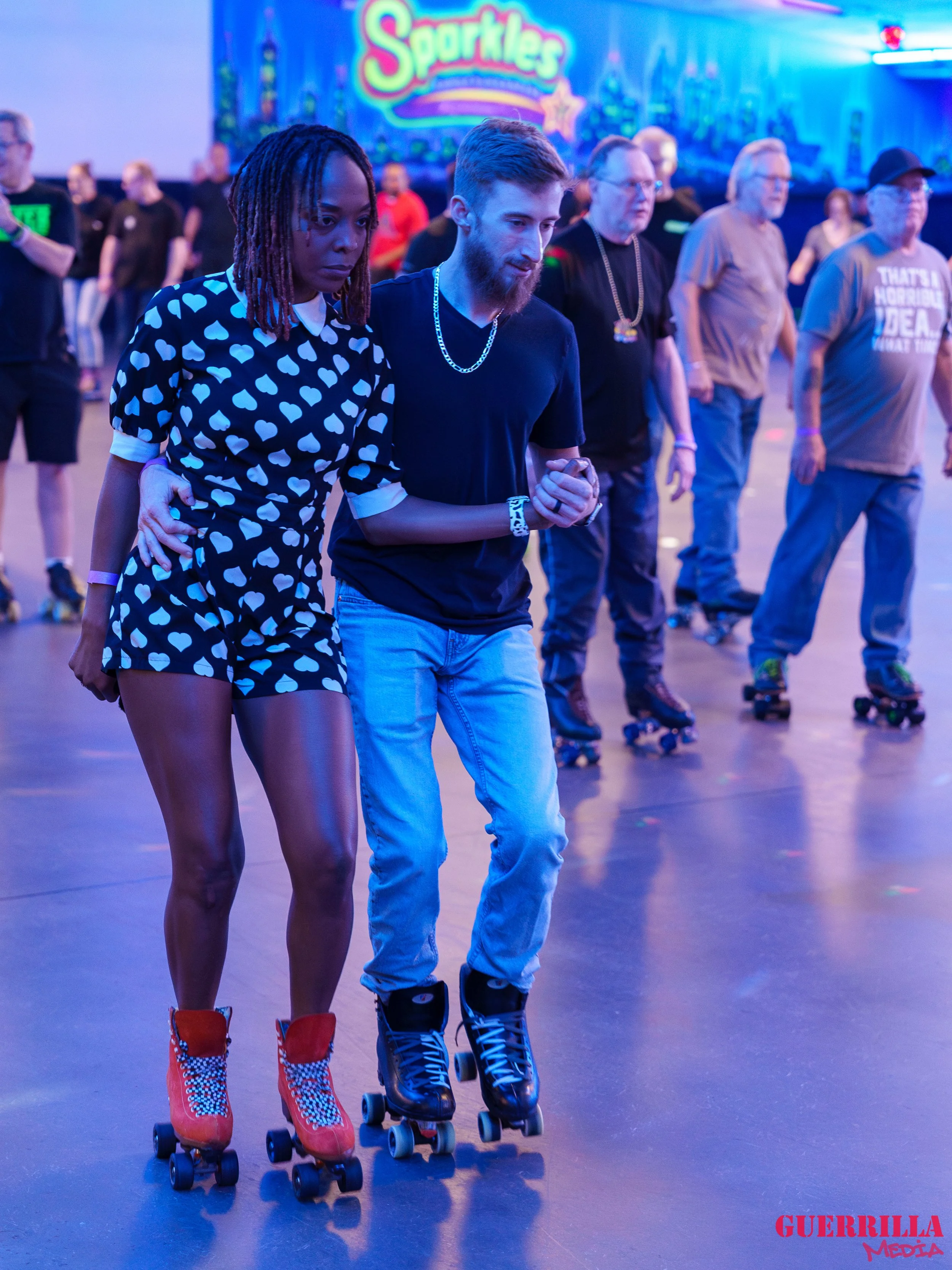 A couple roller skating at an indoor skating rink with other skaters in the background, vibrant lighting, and a colorful wall featuring the word 'Sparkles'.