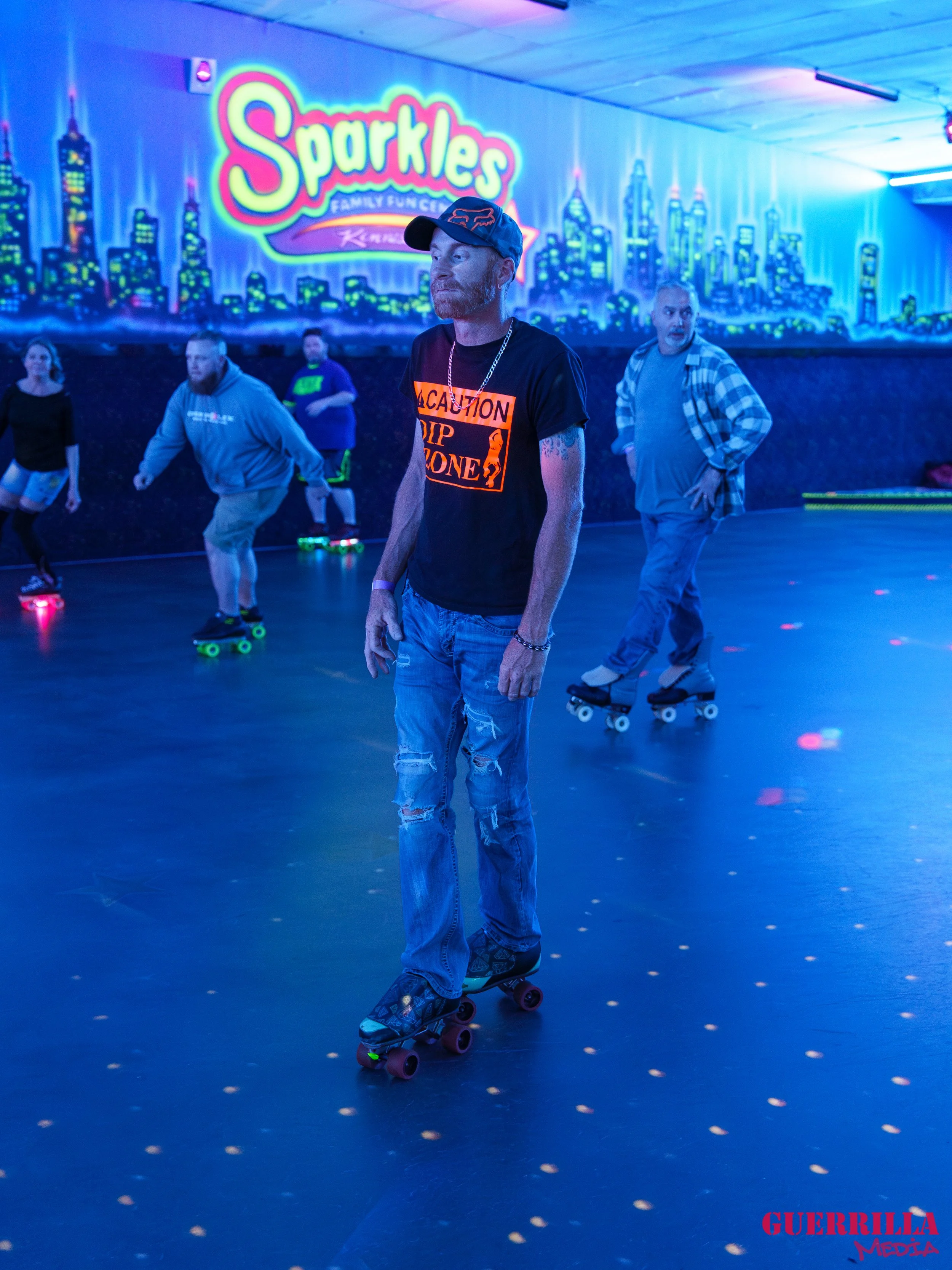 People roller skating in a neon-lit indoor roller rink with a cityscape mural on the wall and a glow sign that reads 'Sparkles Family Fun Center'.