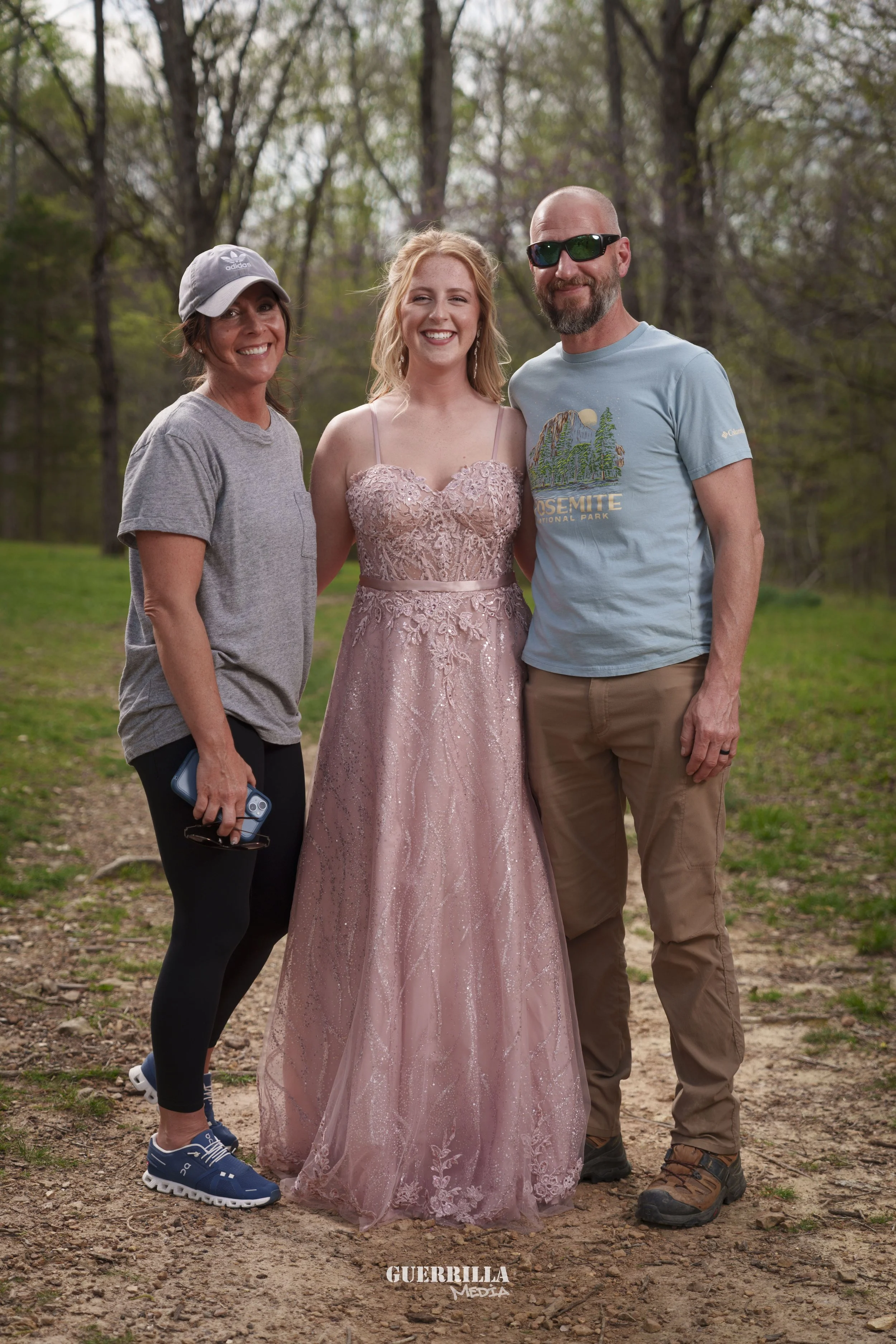 Three people standing outdoors on a dirt path with trees in the background, smiling at the camera. The woman on the left is wearing a gray t-shirt, black leggings, a gray baseball cap, and blue sneakers, holding a phone. The woman in the middle is we