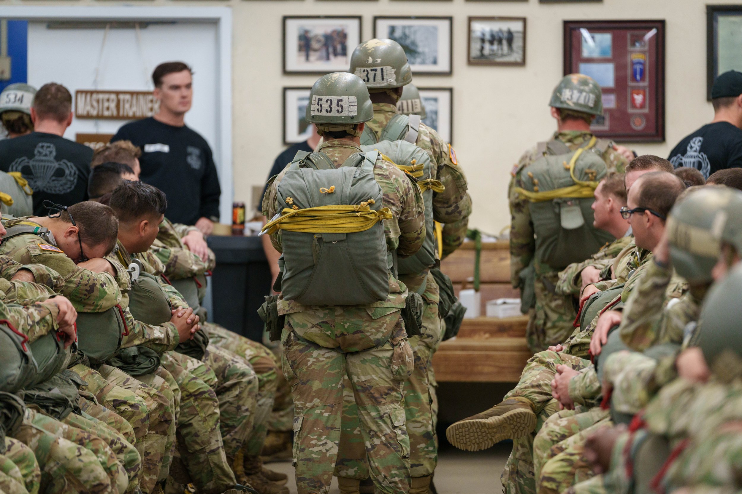 Military personnel in camouflage uniforms and helmets sitting and standing in a room during a training or briefing session, with framed photos on the wall.