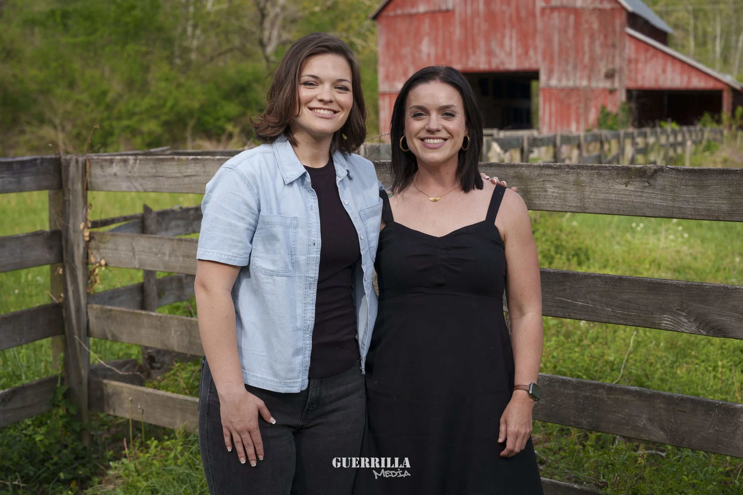 Two women standing outdoors by a wooden fence, smiling at the camera, with a red barn and green trees in the background.