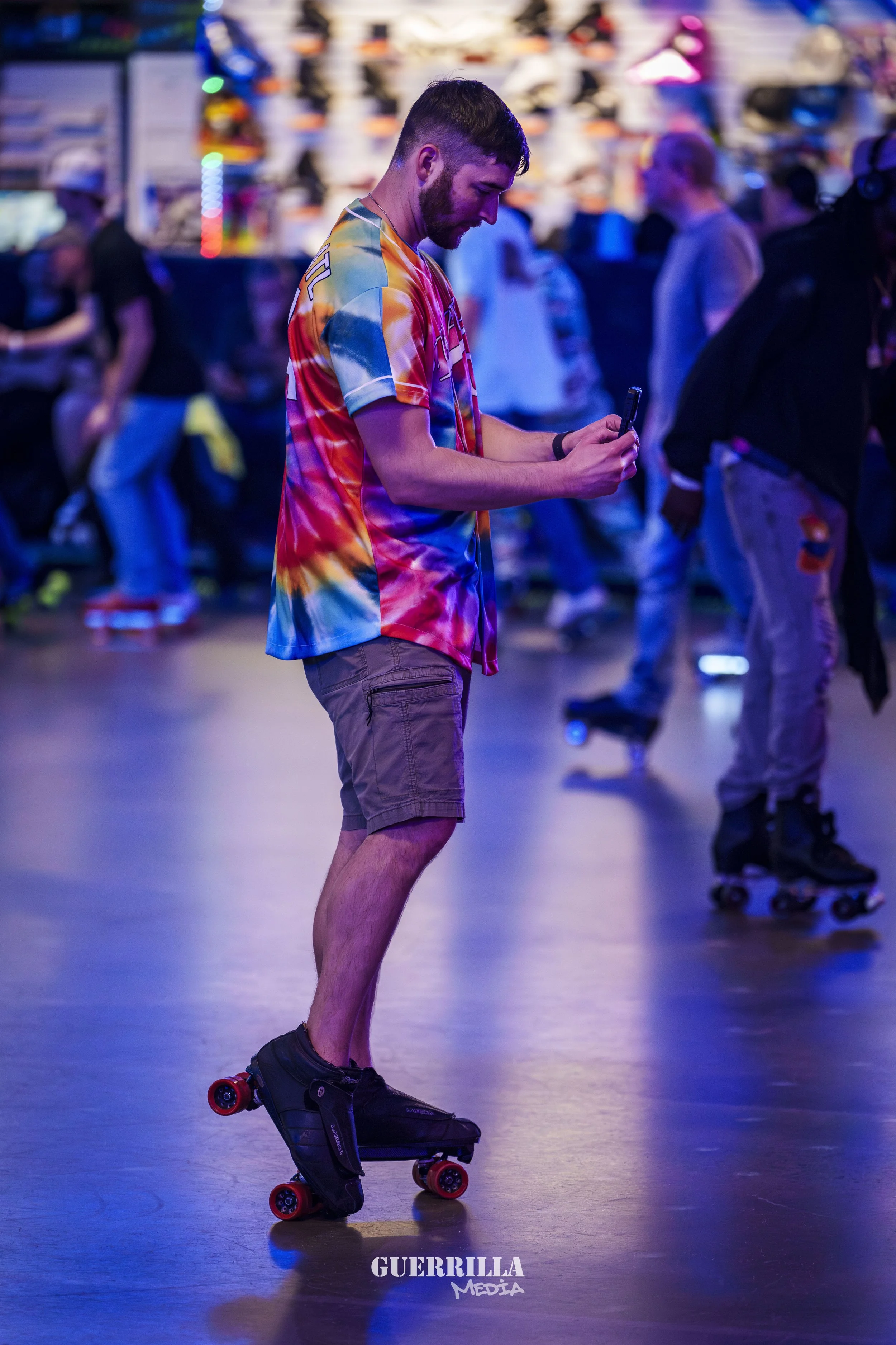 A young man with a beard wearing a colorful tie-dye shirt and khaki shorts is roller skating indoors. He is looking at his phone. There are other people in the background, some also on roller skates, in a lively, colorful setting.