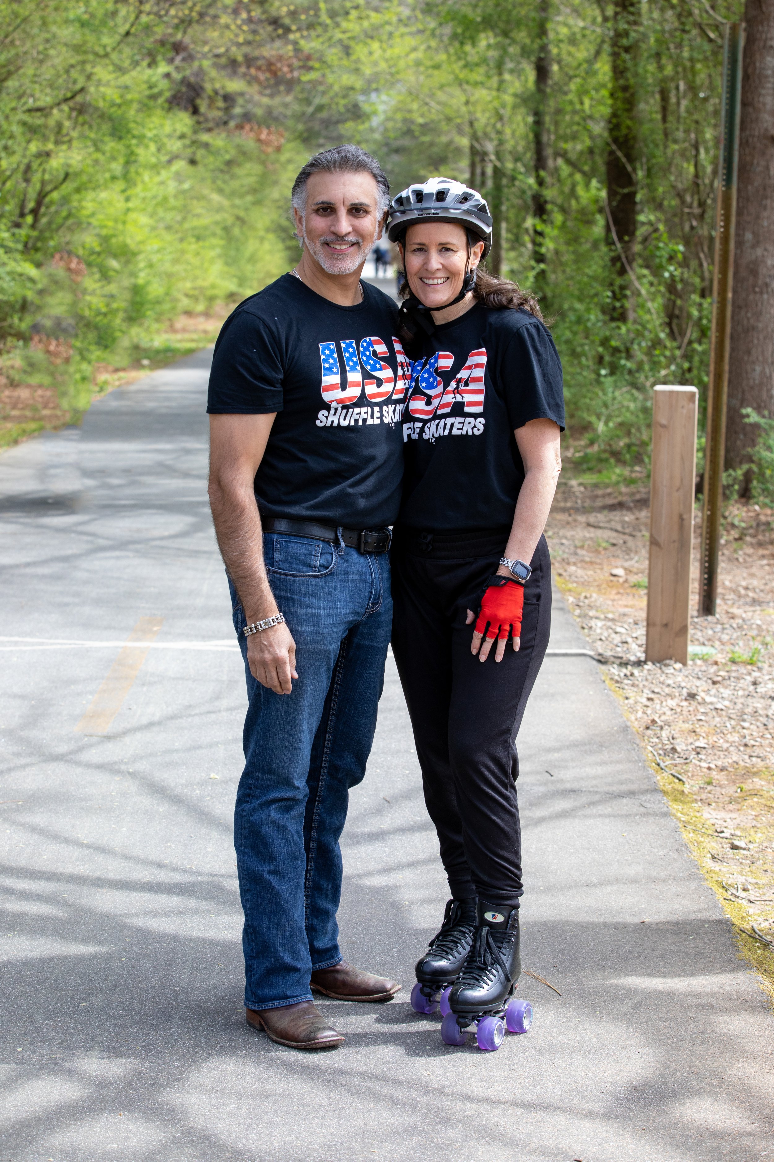 A man and woman standing on a paved path in a wooded area, smiling. The woman is wearing roller skates, a helmet, and a black T-shirt with patriotic designs. The man is dressed casually in jeans and a T-shirt with similar patriotic theme.