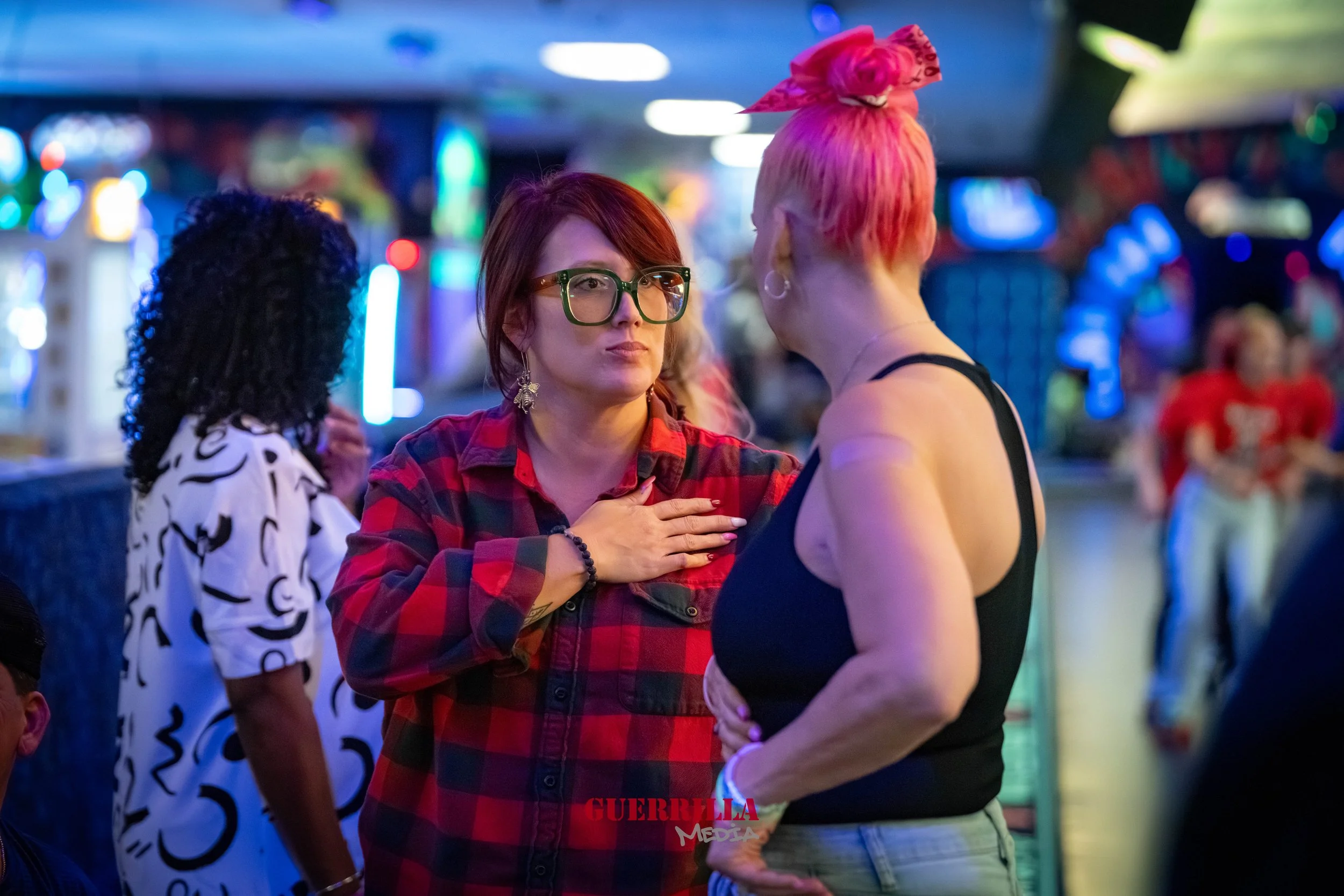 Two women having an intense conversation in a brightly lit arcade, with colorful lights and other people in the background.