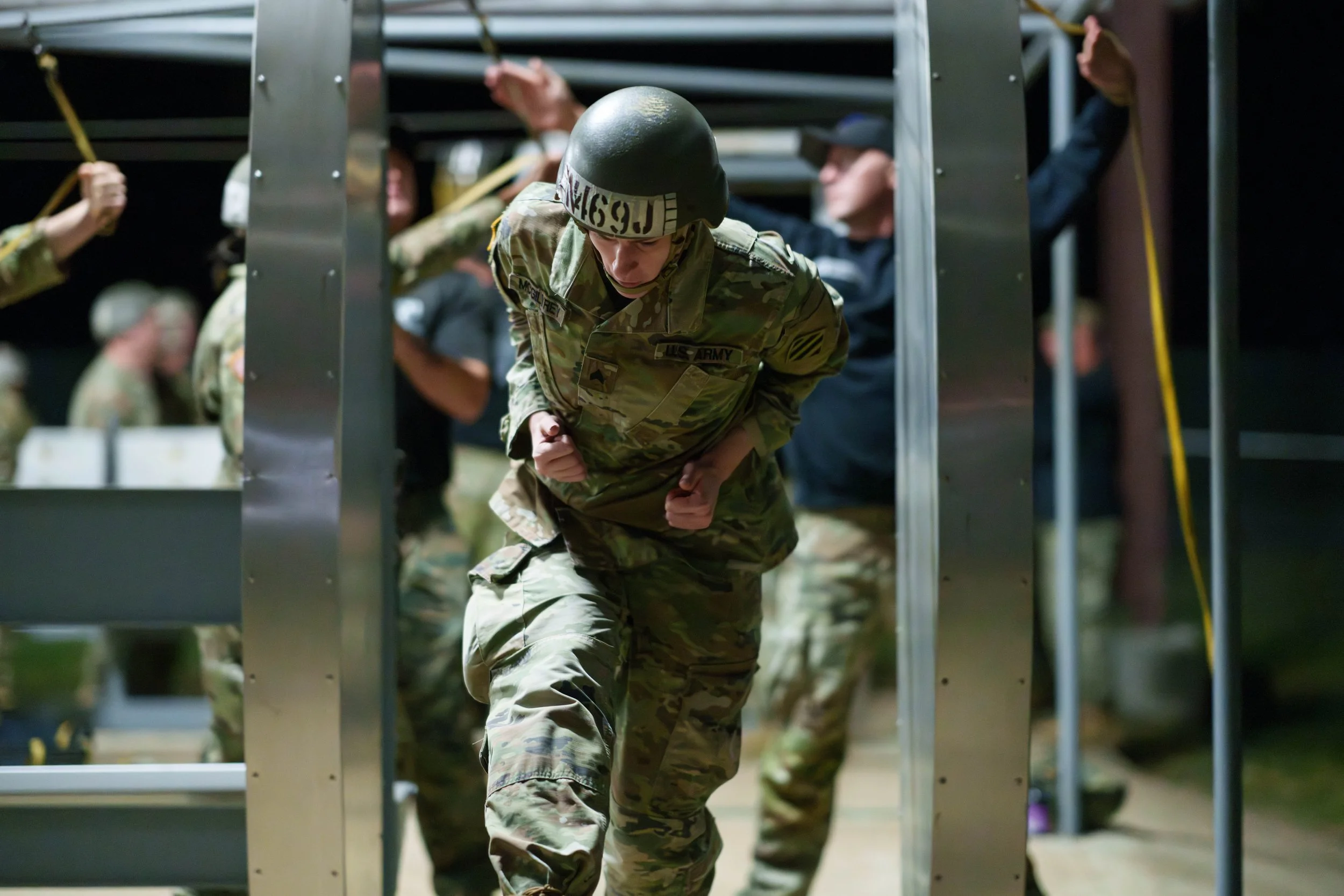 A soldier in full camouflage uniform, helmet, and gear running during a night training exercise surrounded by other soldiers also in camouflage engaging in training activities.