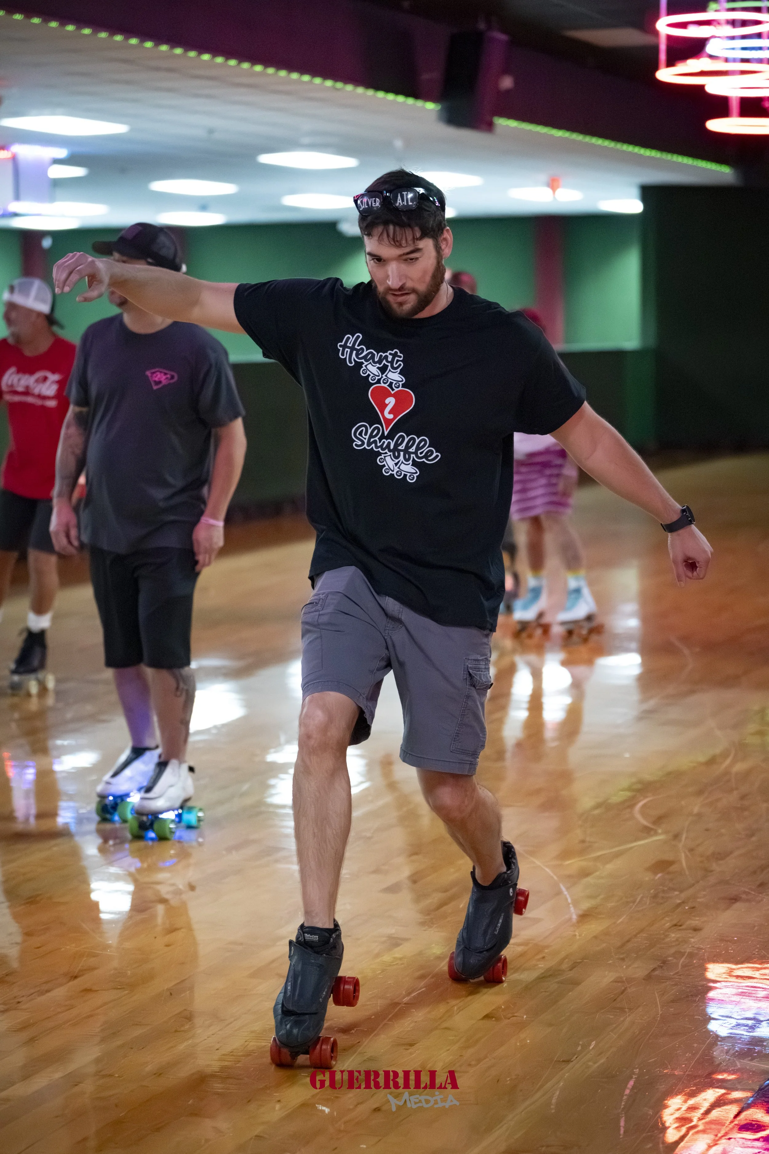 A man skating indoors with other skaters in the background. He wears a black t-shirt, gray shorts, and roller skates with red wheels.