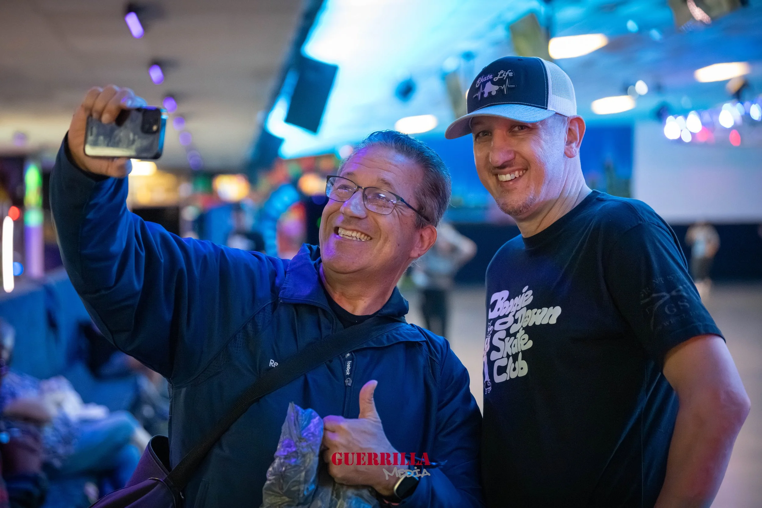 Two men taking a selfie together inside a roller skating rink, with one wearing a blue jacket and glasses, and the other wearing a black T-shirt and a baseball cap. They are smiling and appear to be enjoying the moment.