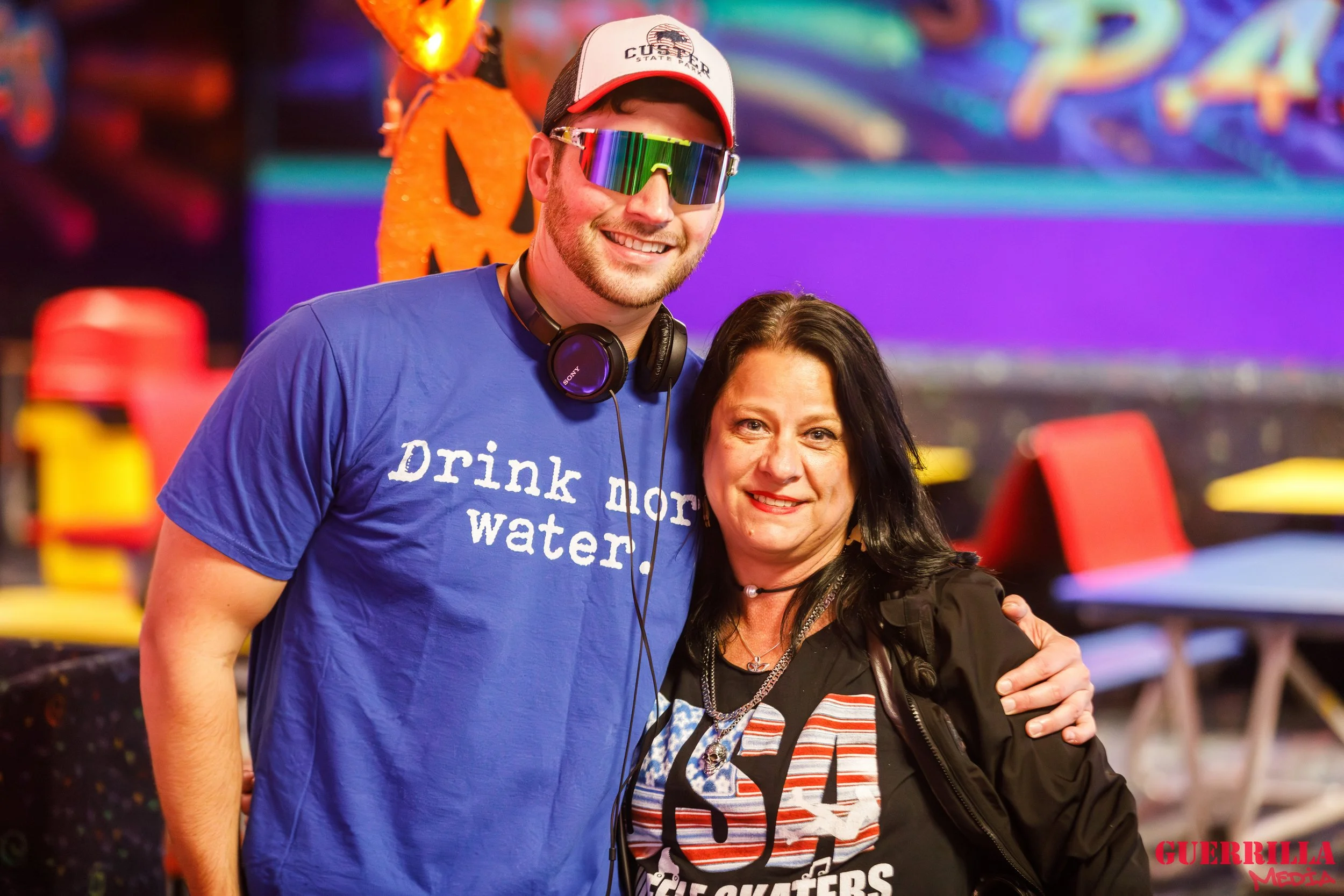 A young man with a beard is wearing a blue t-shirt that says 'Drink more water,' sunglasses with a rainbow neon reflective coating, a baseball cap, and headphones around his neck. He's standing next to a woman with dark hair, wearing a black jacket o
