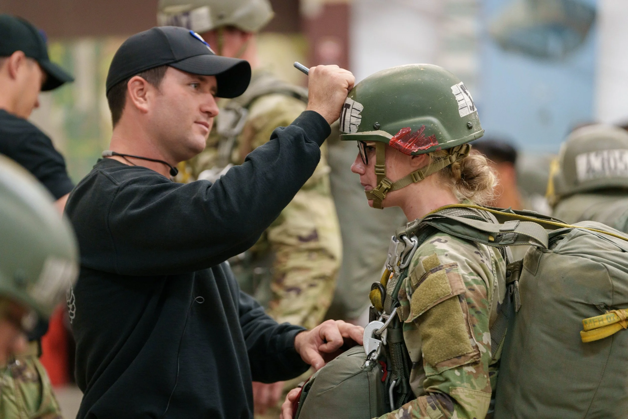 A man helping a female soldier put on her military helmet in a group setting, with other soldiers in the background.