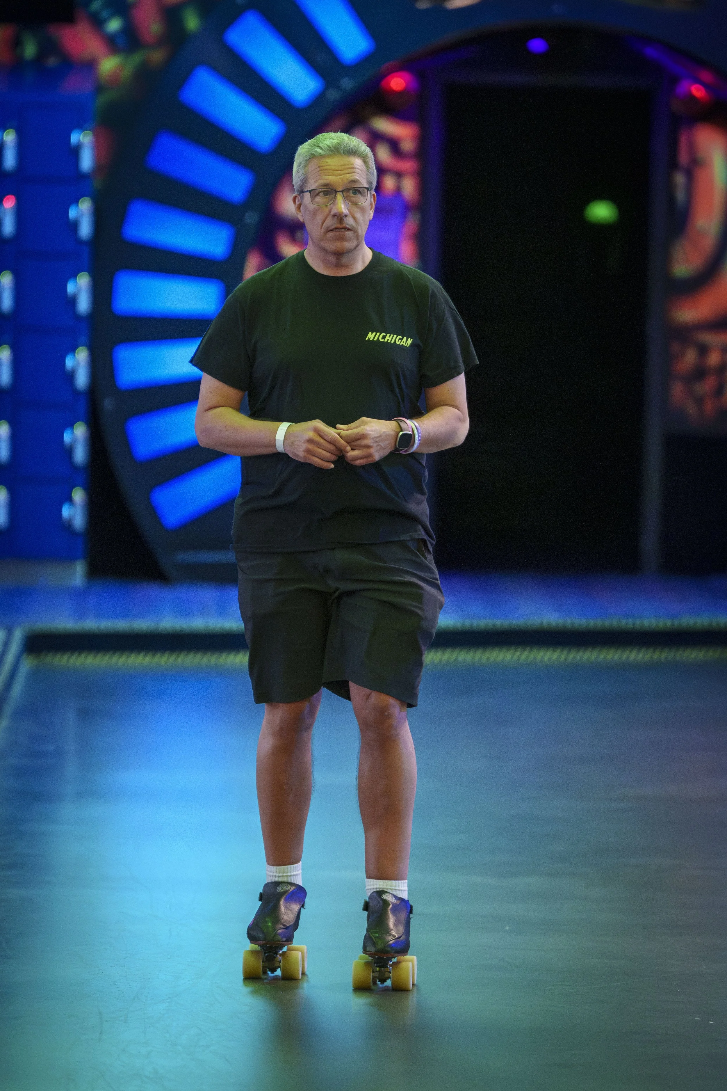 Man wearing a black T-shirt with 'Michigan' printed on it, black shorts, and roller skates stands on a roller rink.
