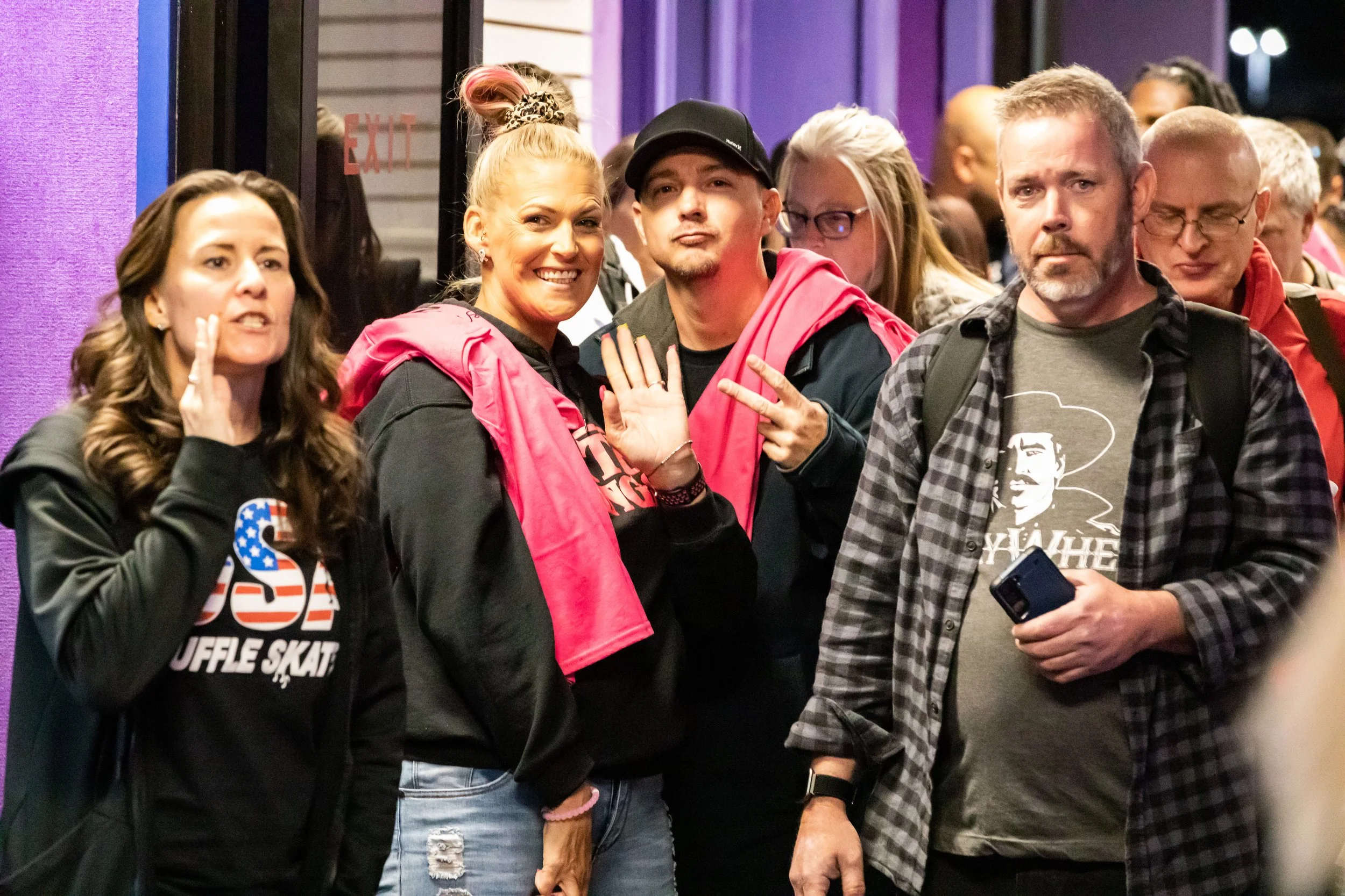 Group of five people standing together at an event, with some looking at the camera and others engaged in conversation, in front of a purple wall.