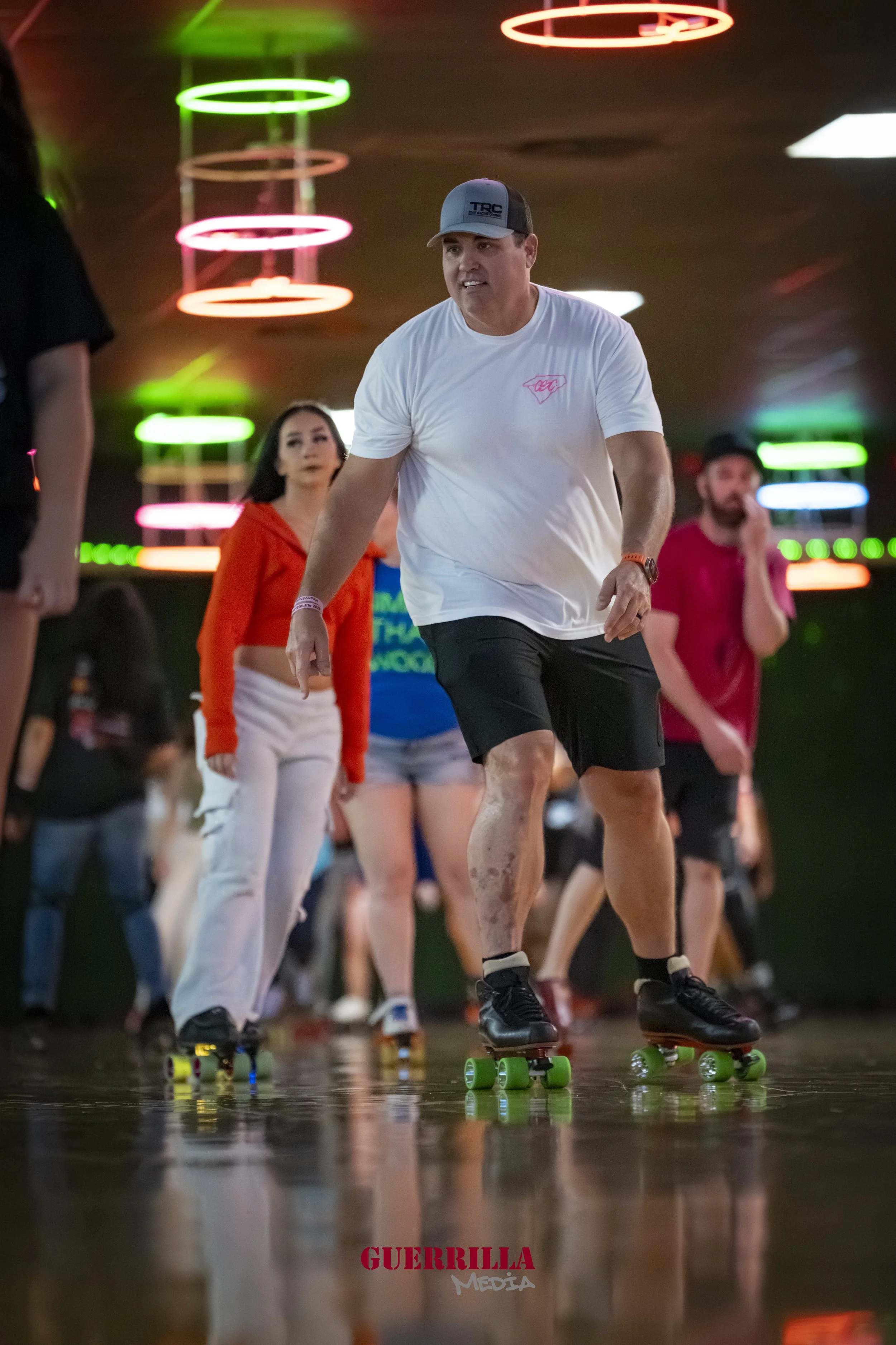 People roller skating indoors with neon lights hanging from the ceiling, focusing on a man in a white T-shirt and black shorts.
