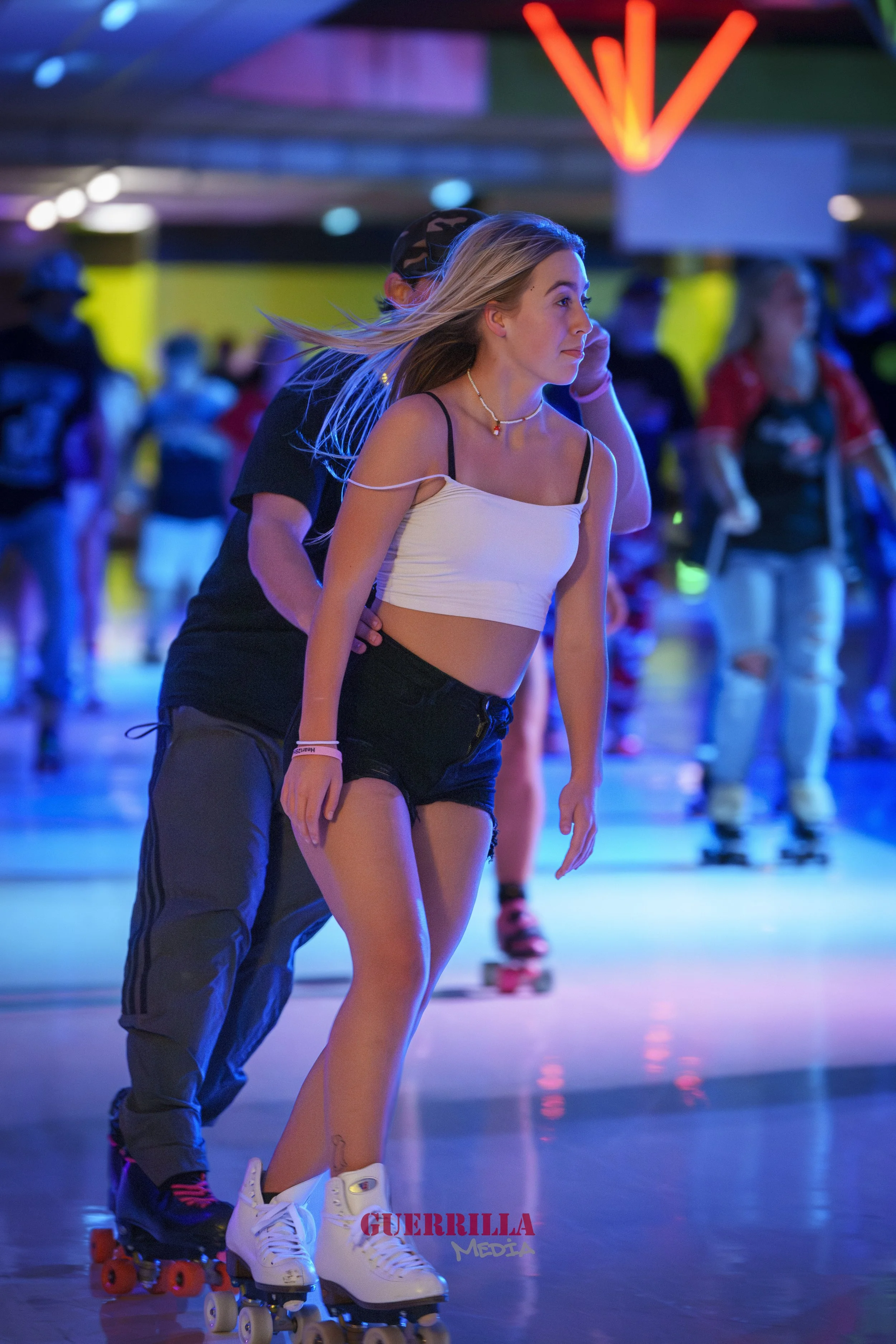 A young woman roller skating in an indoor skating rink with colored neon lights, with a man behind her guiding her. Other skaters are visible in the background.