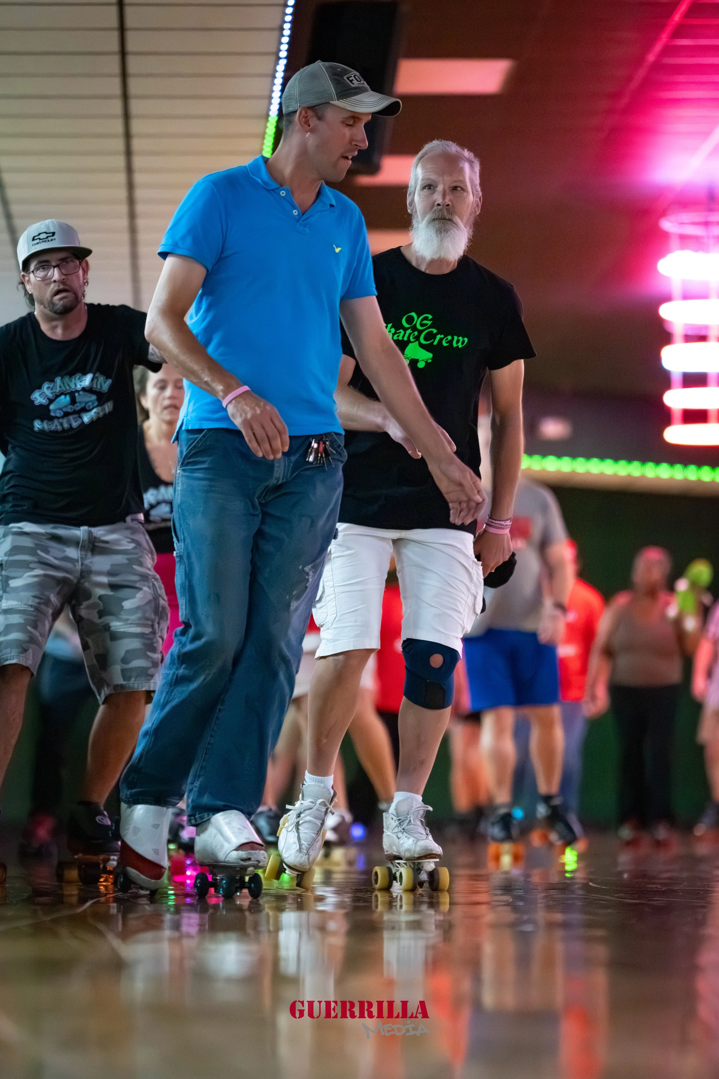 People roller skating indoors; one man in a blue shirt and cap holding hands with a woman wearing white shorts and a knee brace, surrounded by others skating; colorful neon lights in the background.