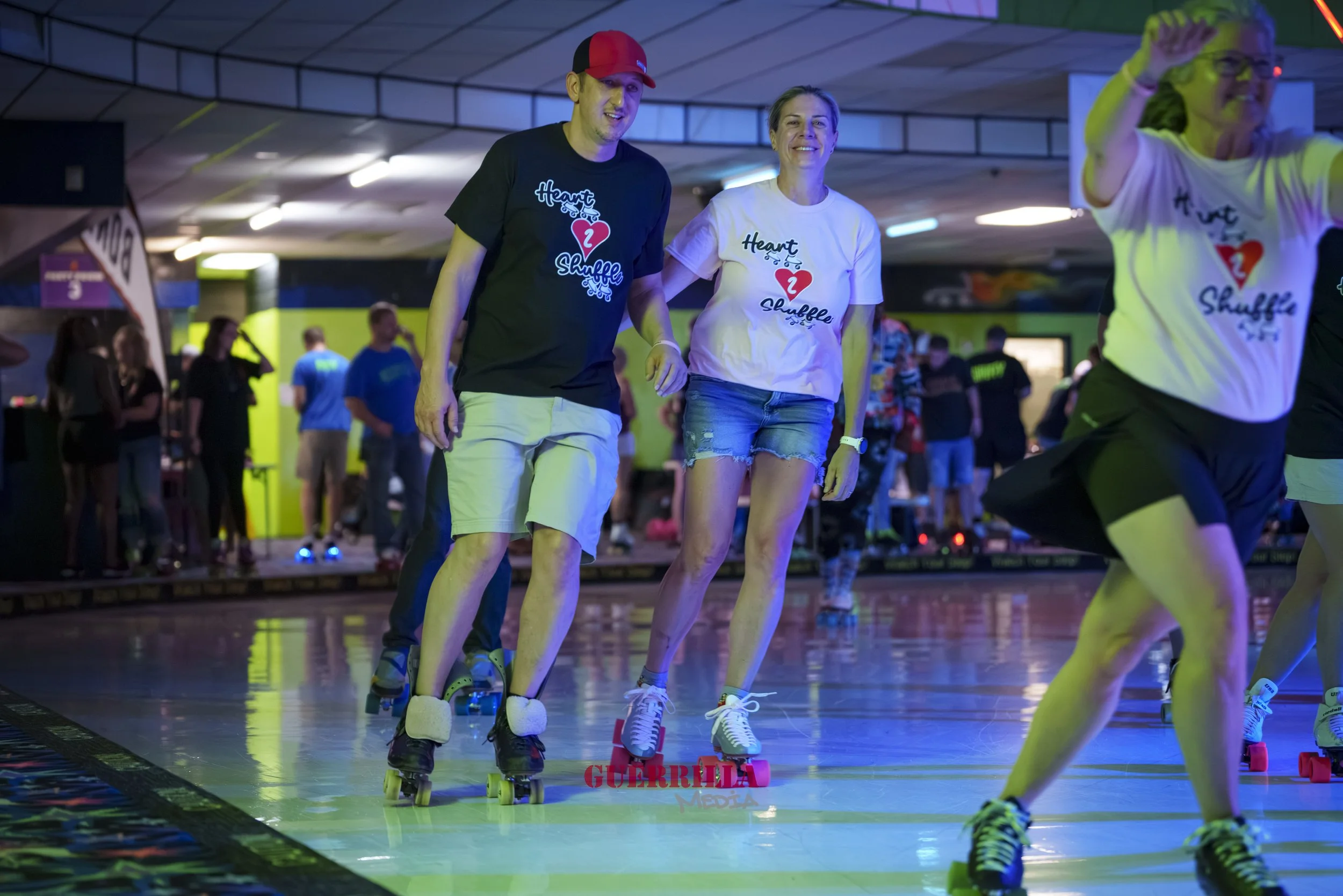 People roller skating at an indoor rink, wearing matching t-shirts that say 'Heart Shufflez' with a red heart graphic. The rink has colored lighting and a crowd of spectators in the background.