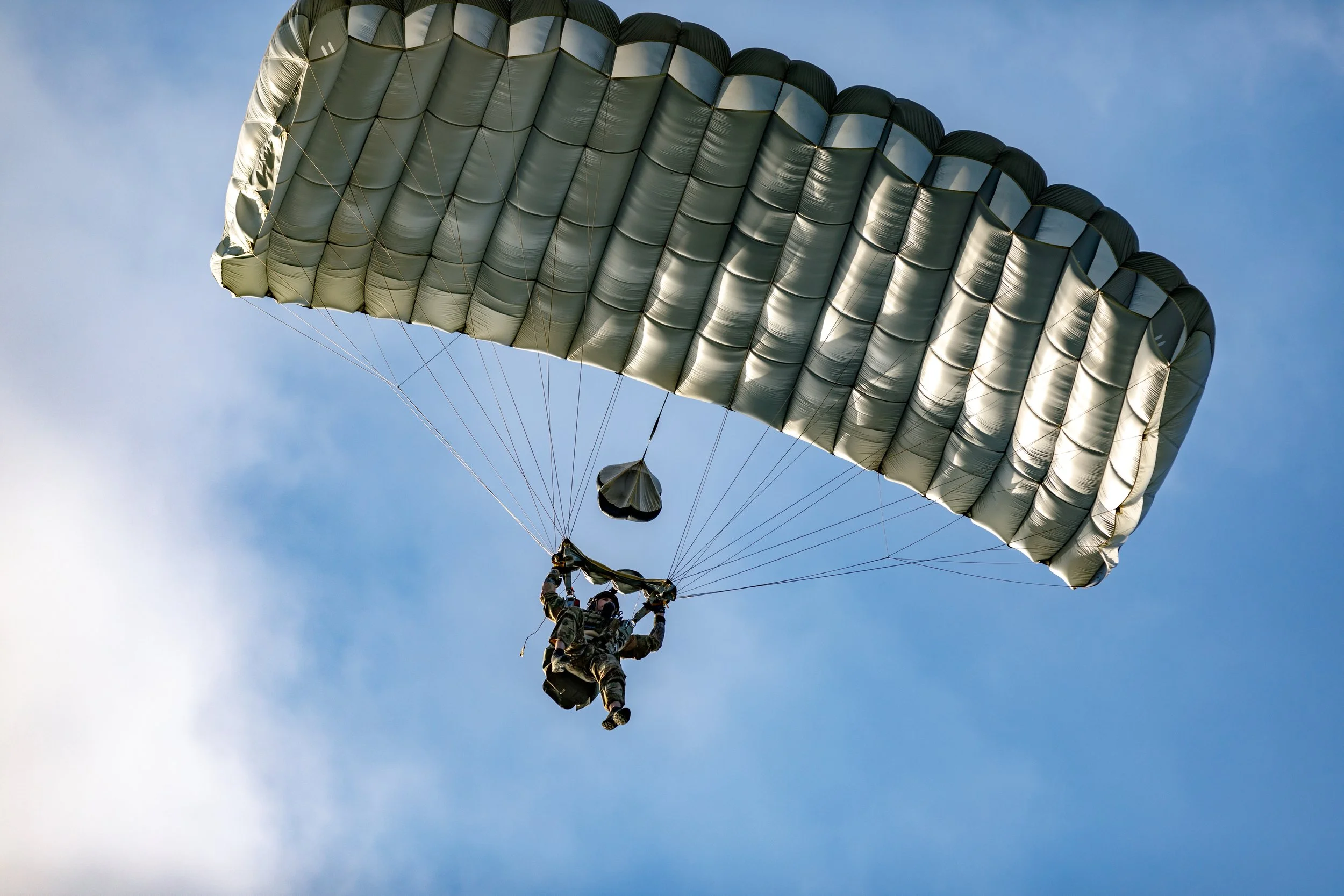 A person in military gear parachuting with a silver and black parachute in a clear blue sky.