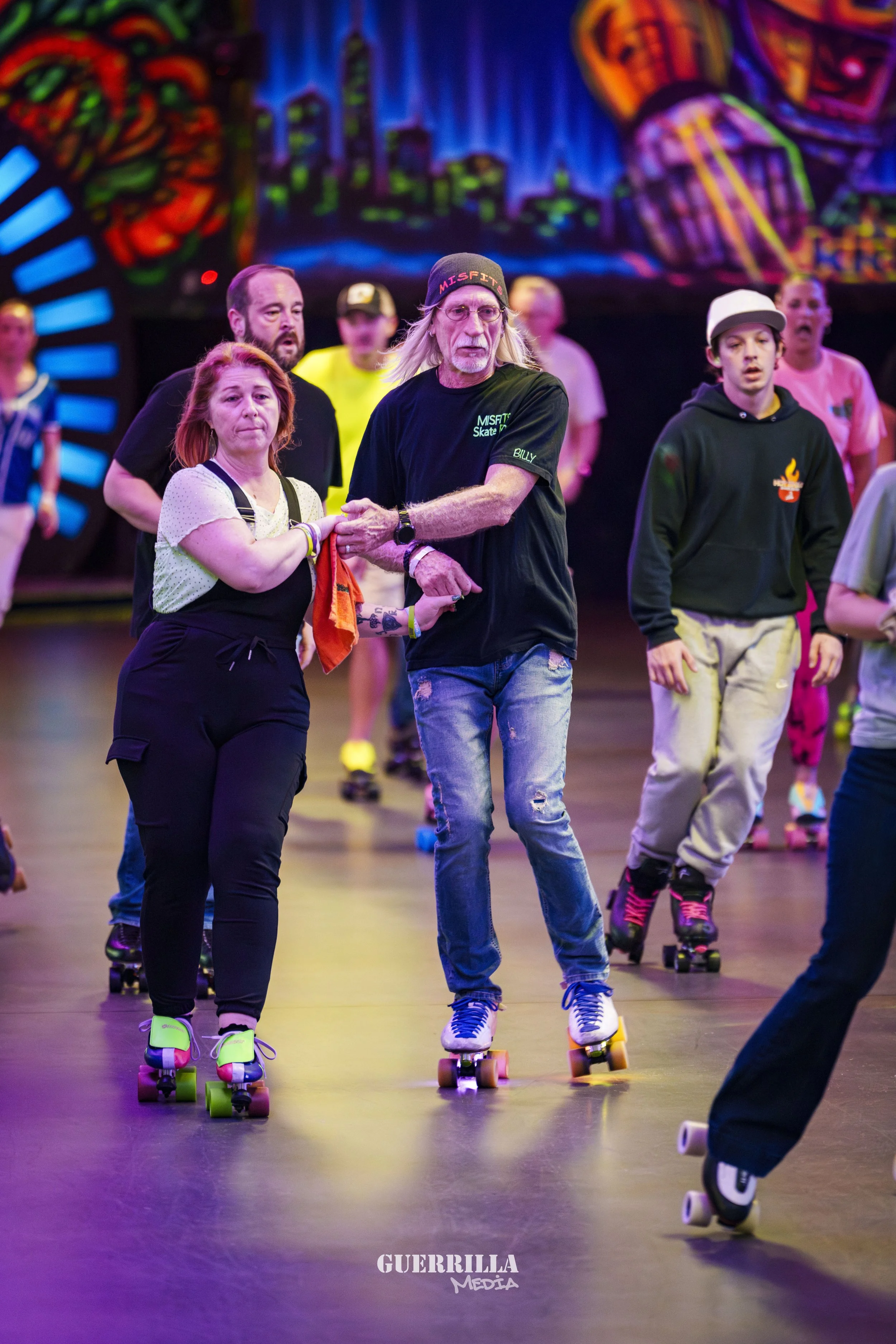 People roller skating in a brightly lit indoor space with colorful mural art on the walls. A man with long hair and a beanie holds hands with a woman as they roller skate. Several other skaters are visible in the background.