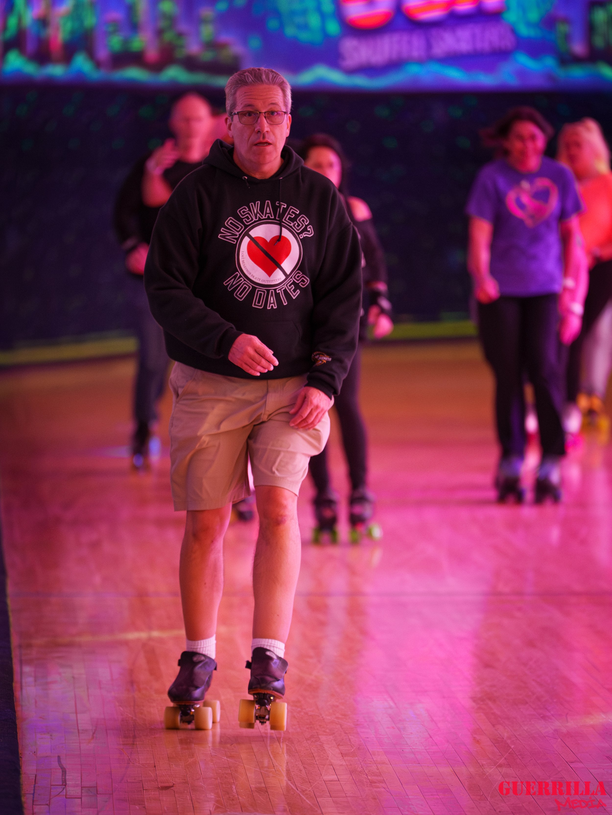 A man roller skating on a wooden floor at an indoor rink, wearing a black hoodie with a heart and the words 'No Skates? No Dates,' and beige shorts. Behind him, multiple people are skating together under colorful lights.