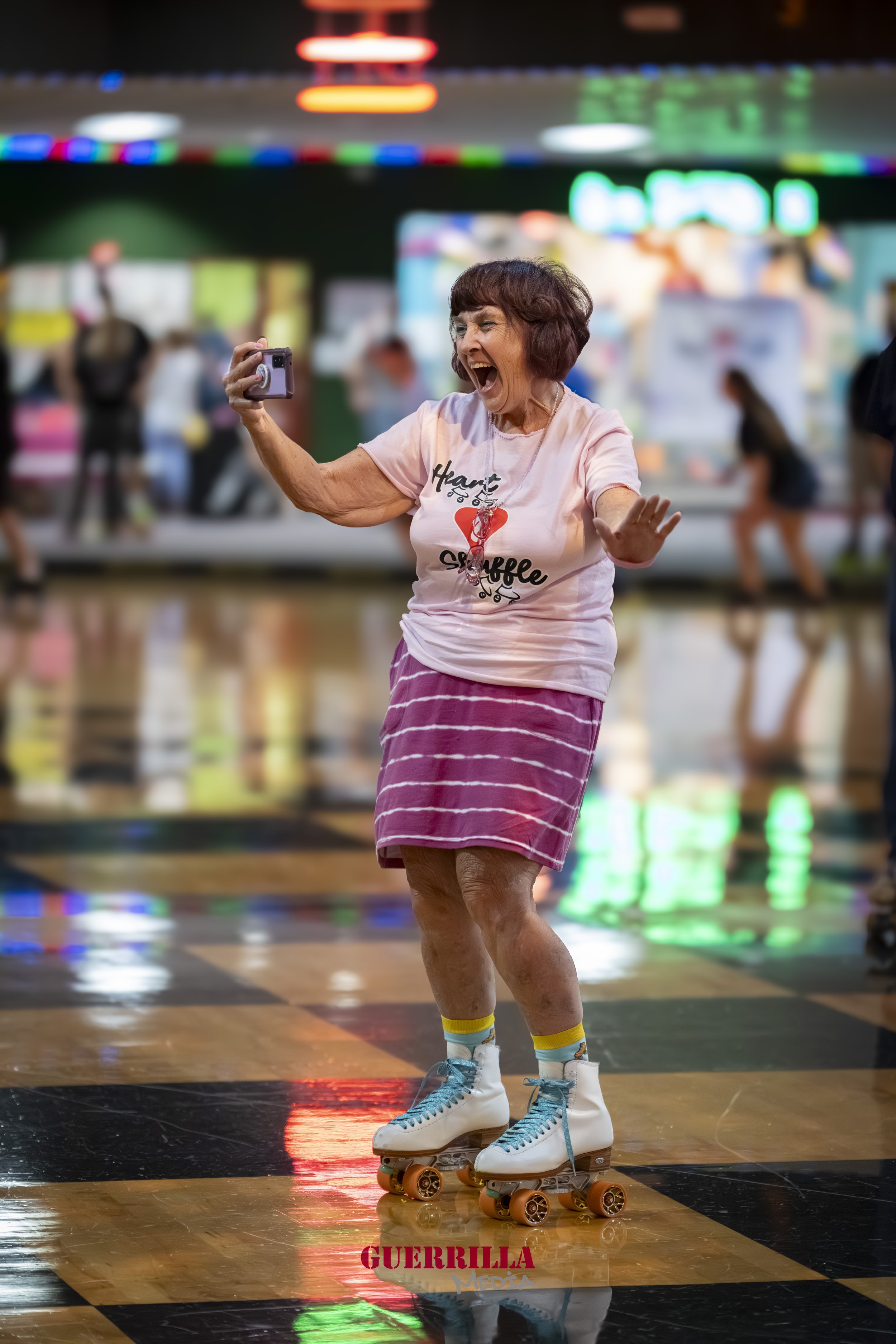 An elderly woman skating indoors on roller skates with a big smile, holding a camera, wearing a pink skirt, a white t-shirt, and colorful socks, with a lively background of neon lights and people.