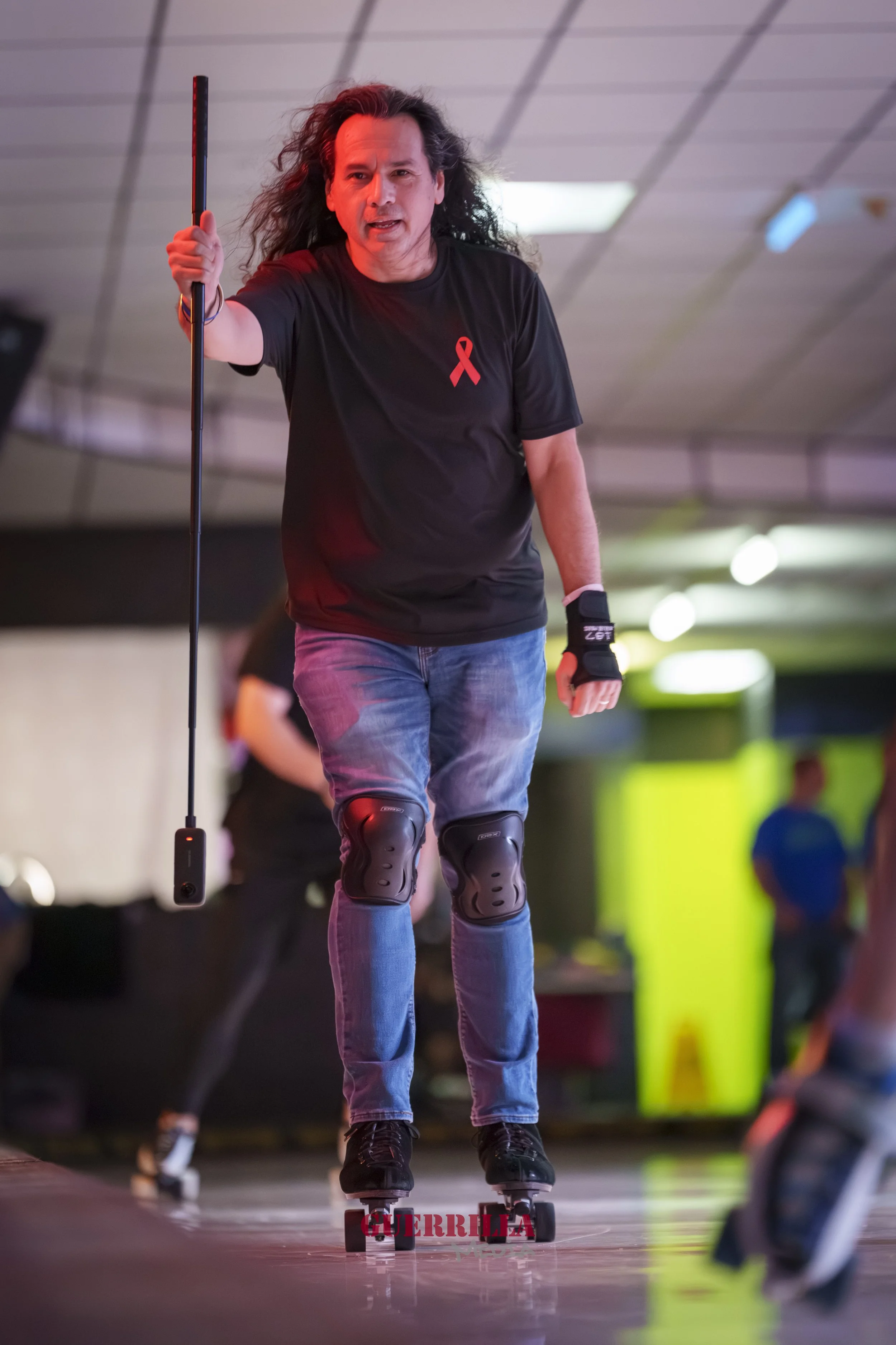 Man roller skating indoors, wearing knee pads, wrist guards, a black t-shirt with a red ribbon, and holding a skate tool in his right hand.