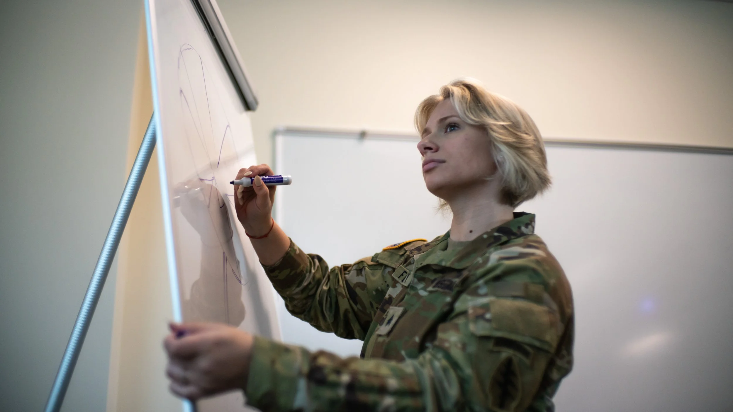 Female soldier in camouflage uniform writing on a whiteboard with a marker.