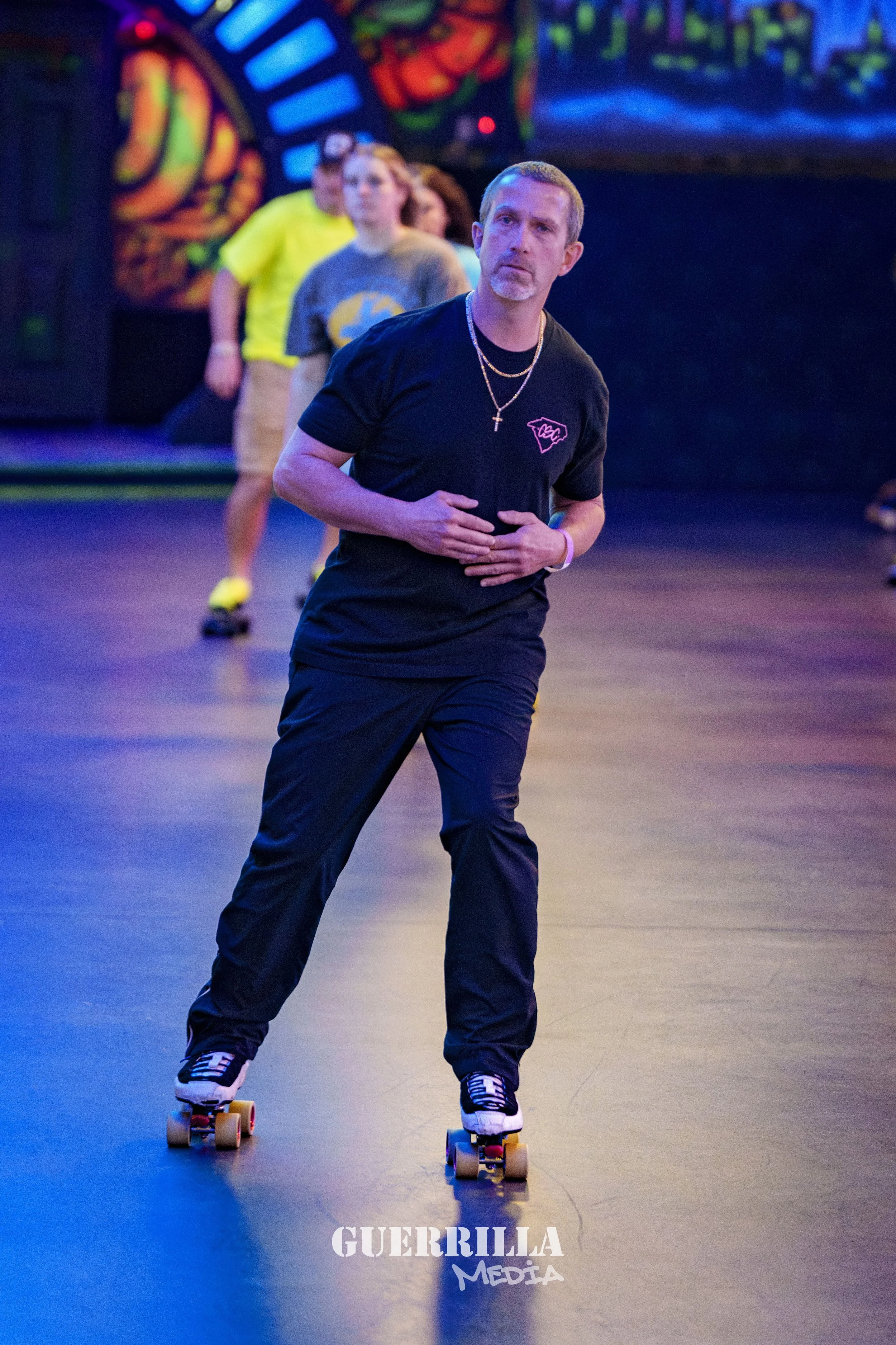 A man with short hair and a goatee wearing a black t-shirt, black pants, and roller skates, standing on a roller rink with his hands on his stomach. Behind him, colorful neon lights and other skaters are visible.