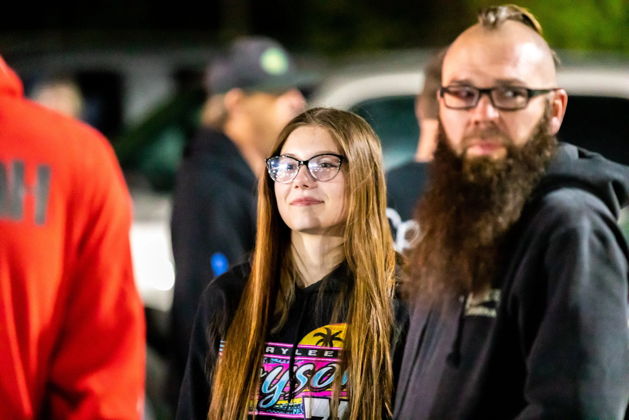Two people, a woman with long red hair and glasses, and a man with a long beard and glasses, are standing outdoors at night. The woman is wearing a black hoodie with a colorful graphic design, and the man is wearing a black jacket. There are several 