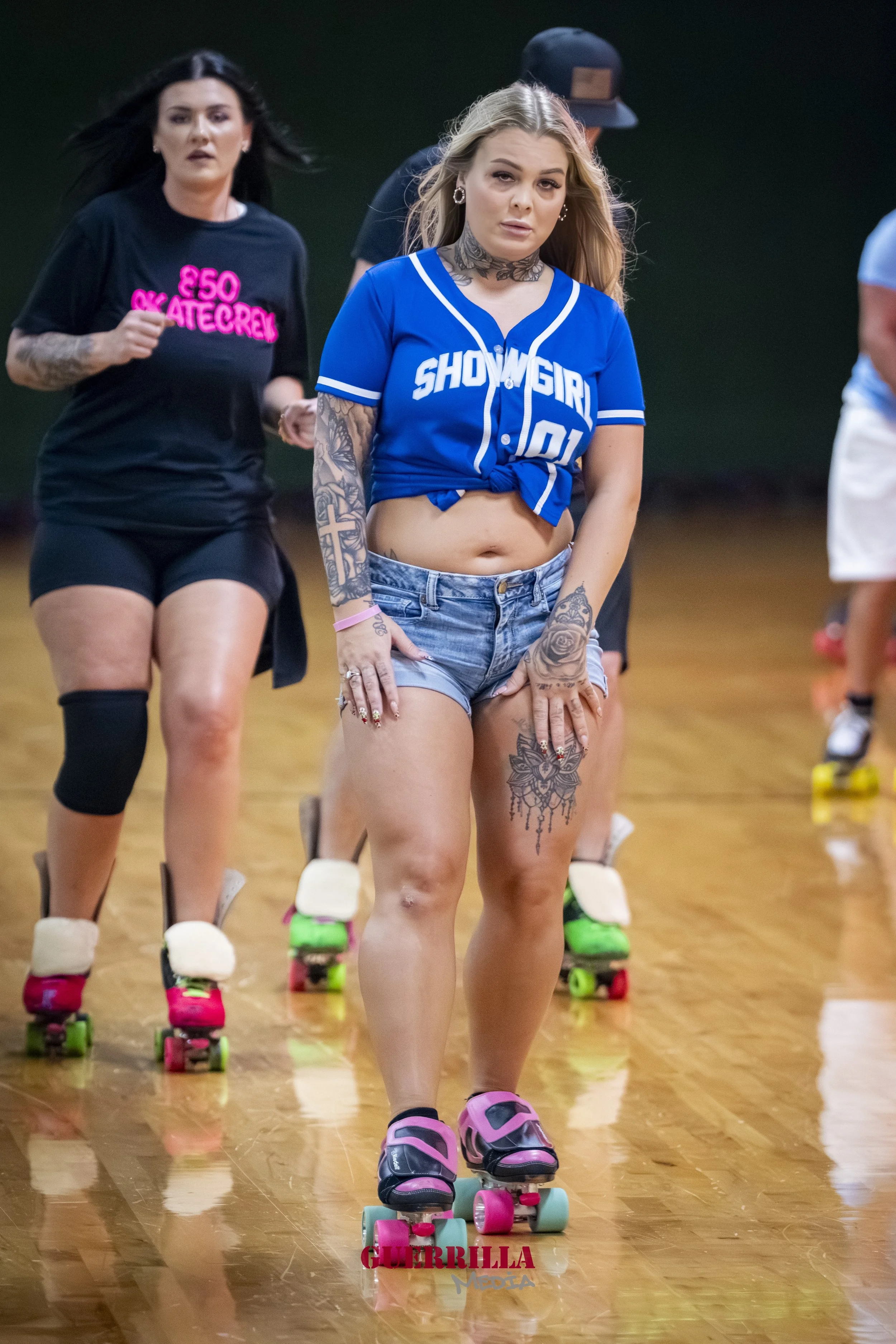 Two women roller skating indoors, one wearing a blue sporty shirt and denim shorts, the other in a black t-shirt with pink text, both on pink roller skates.