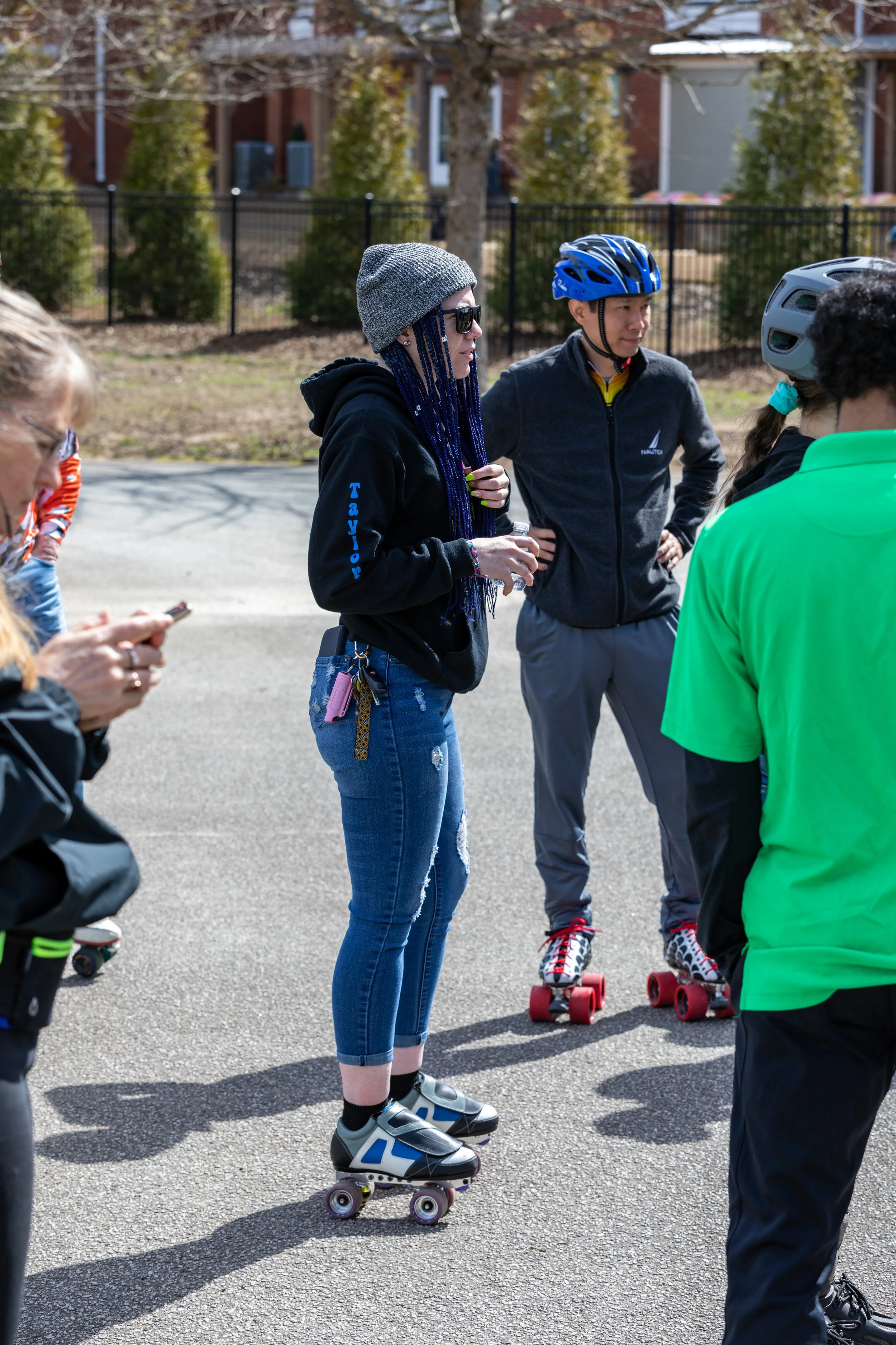 A group of people wearing helmets and roller skates standing outside on a paved surface, with a woman in the center talking and others listening.