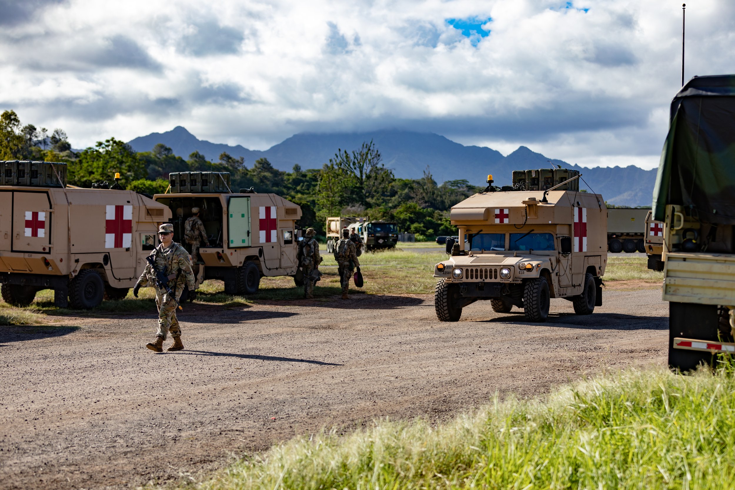 Military personnel and vehicles with Red Cross symbols in a rural area with mountains and cloudy sky in the background.