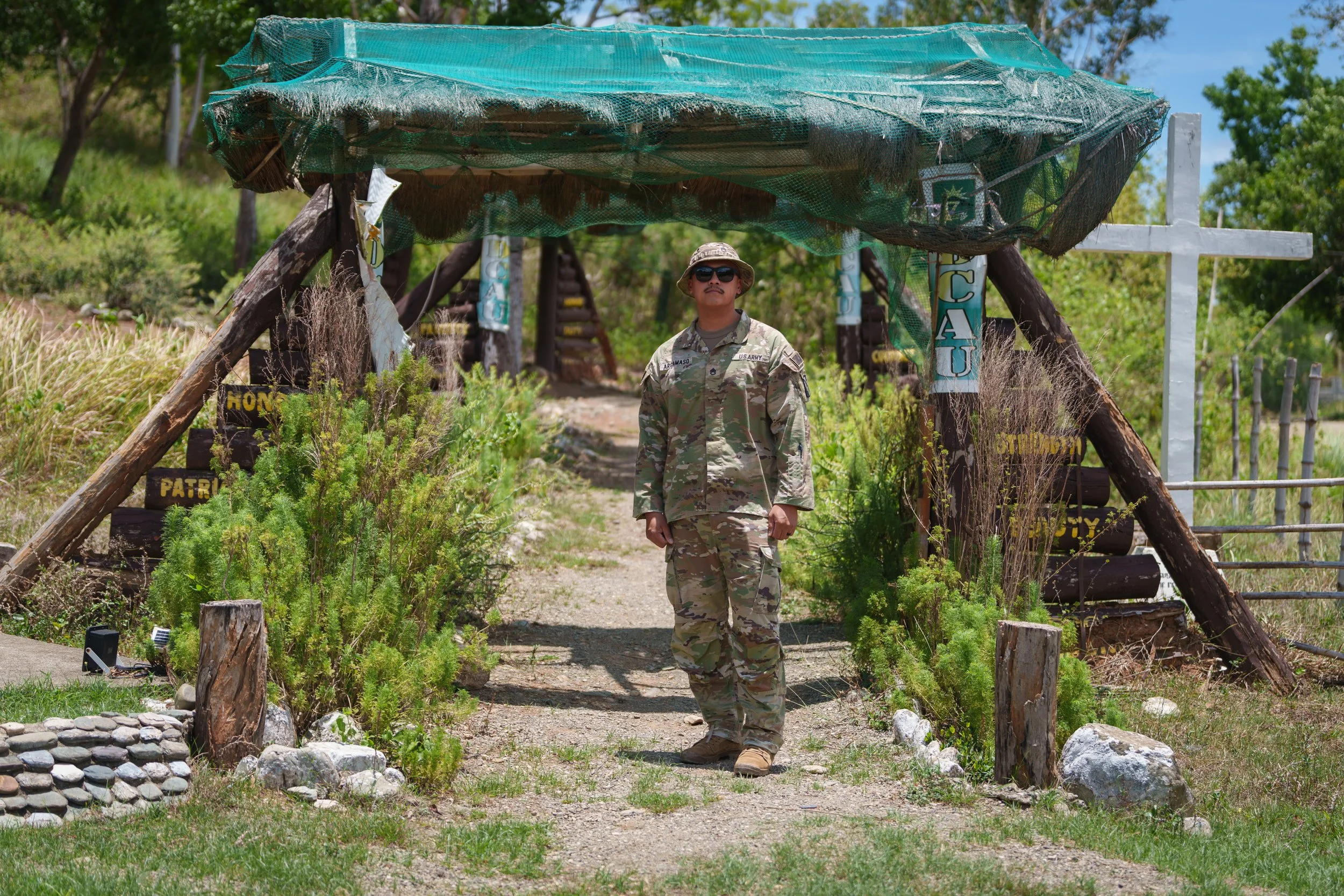A woman in military camouflage standing under a makeshift shelter with a cross and horseback riding signs, surrounded by greenery.