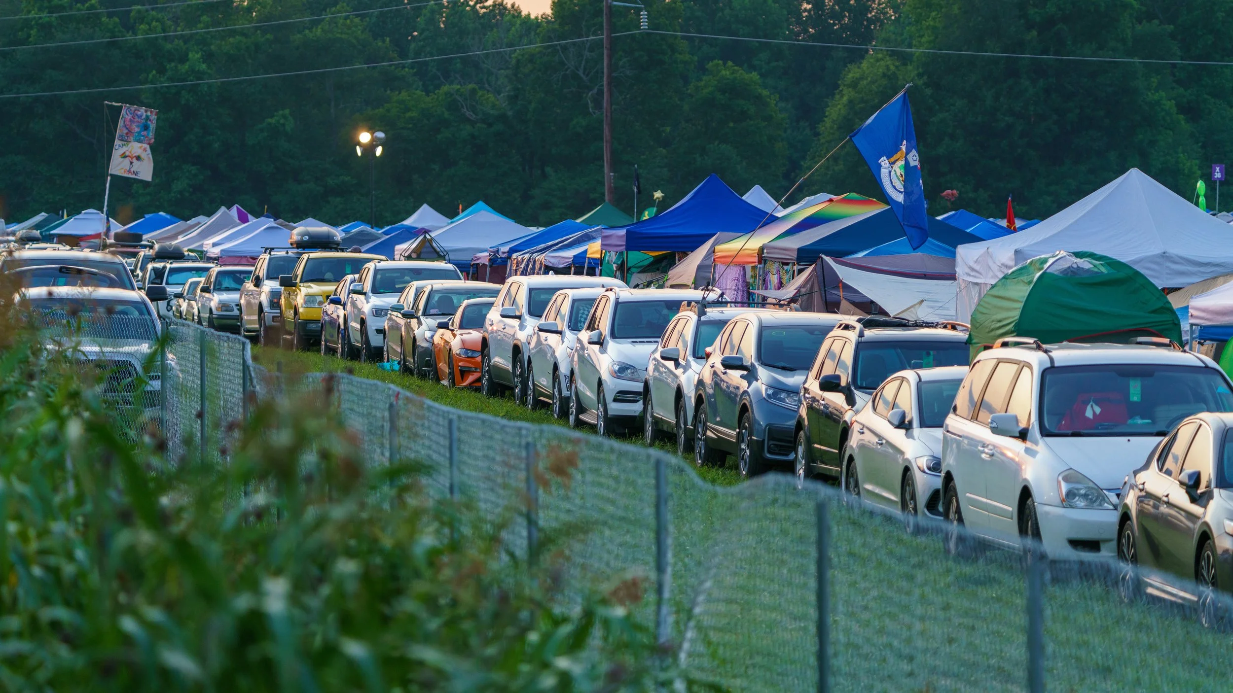 Cars parked in a row alongside camping tents at an outdoor event with trees in the background.