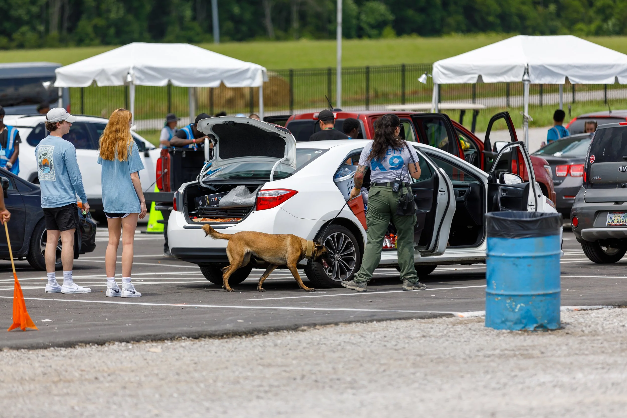 People in a parking lot with a white car, some opening doors, a woman with a Walkie Talkie, a dog on a leash, and tents in the background.
