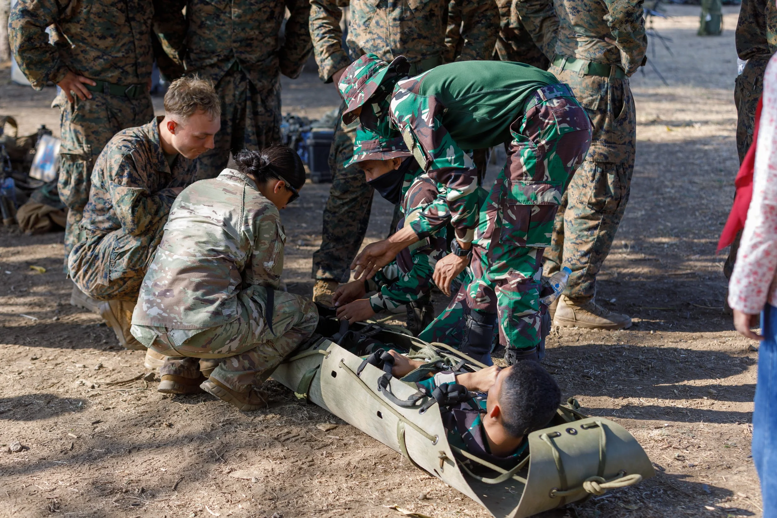 Military personnel assisting a soldier into a stretcher during a training exercise outdoors.