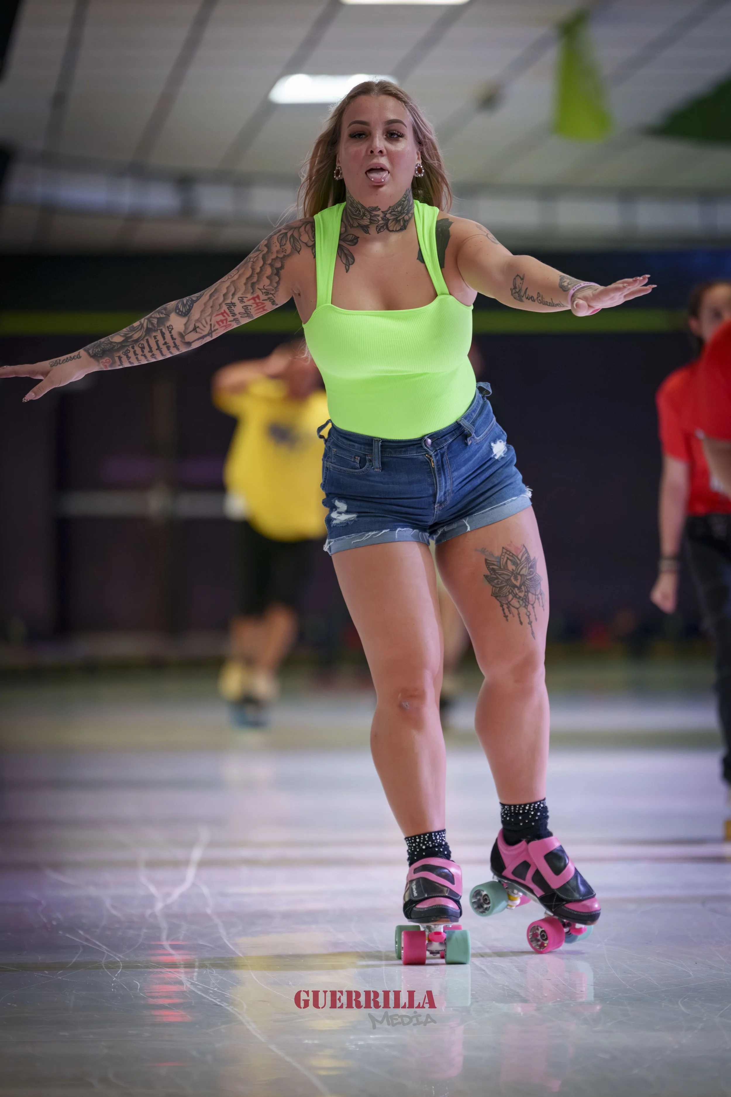 A woman roller skating indoors, wearing a neon green top, denim shorts, and black socks with pink roller skates, with her arms extended as she balances.