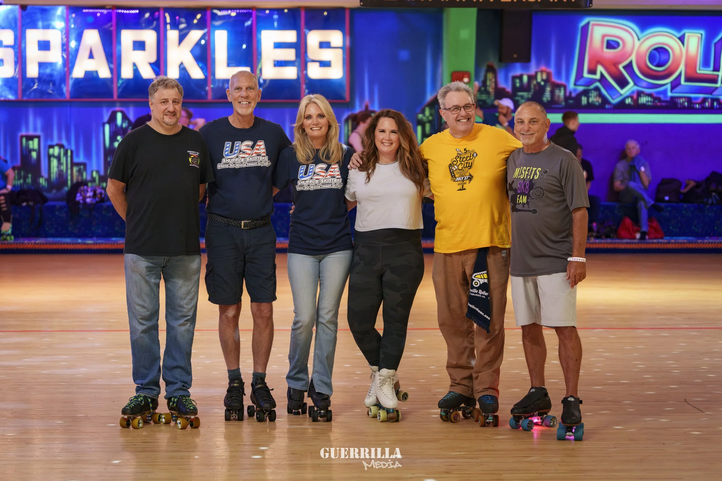 Group of seven people skating on an indoor roller rink with colorful lights and neon signs in the background, smiling and posing for the photo.