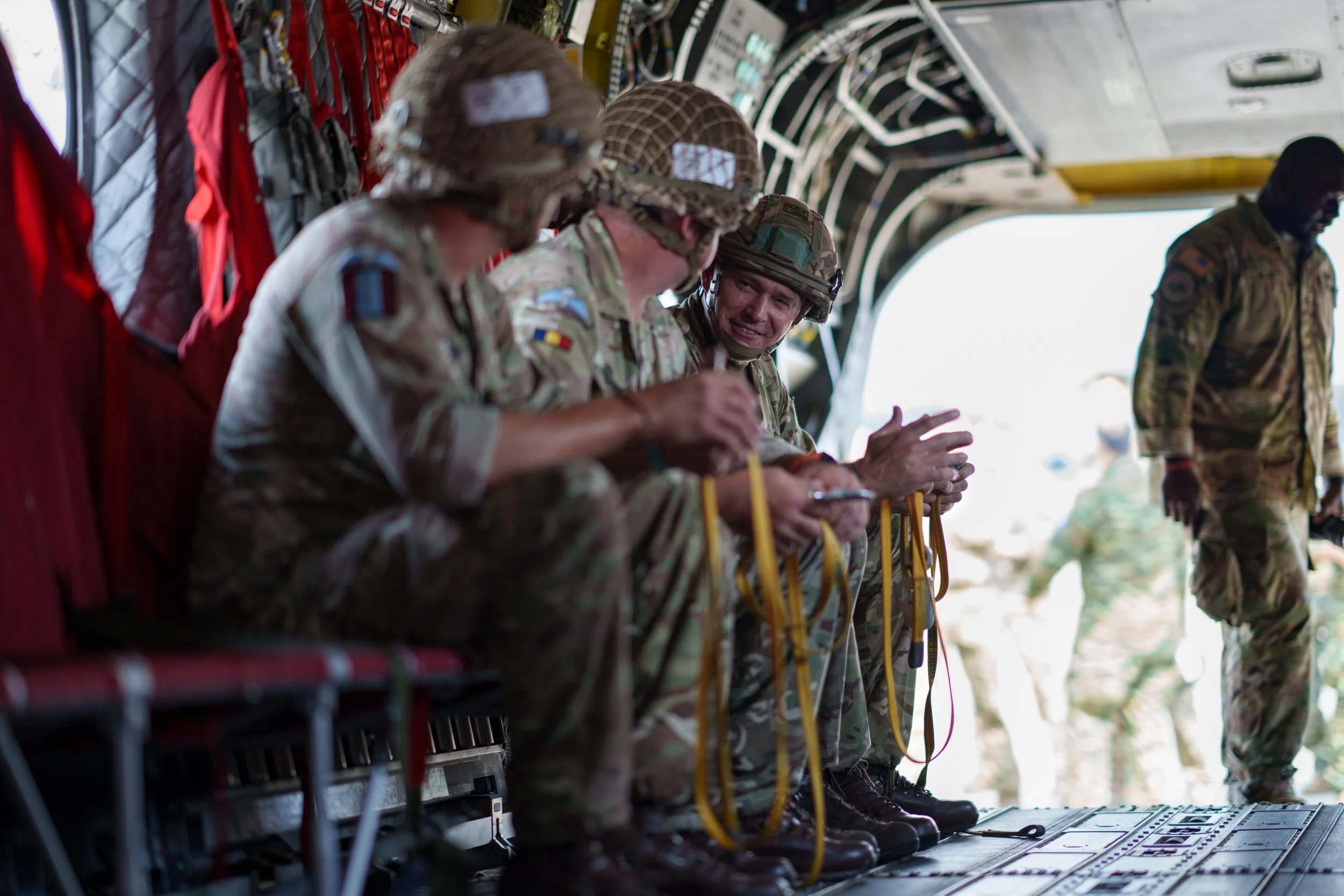 Military personnel in camouflage uniforms sitting and preparing equipment inside a military aircraft, with some standing outside.