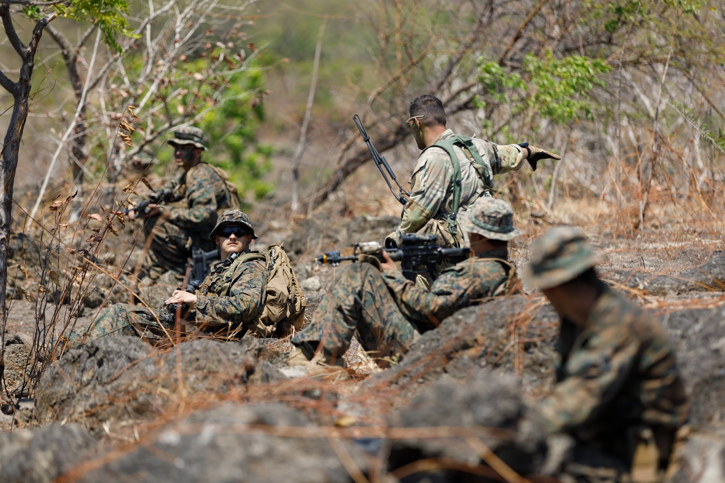 Five soldiers in camouflage uniforms and gear resting and talking in a rocky, wooded outdoor area.