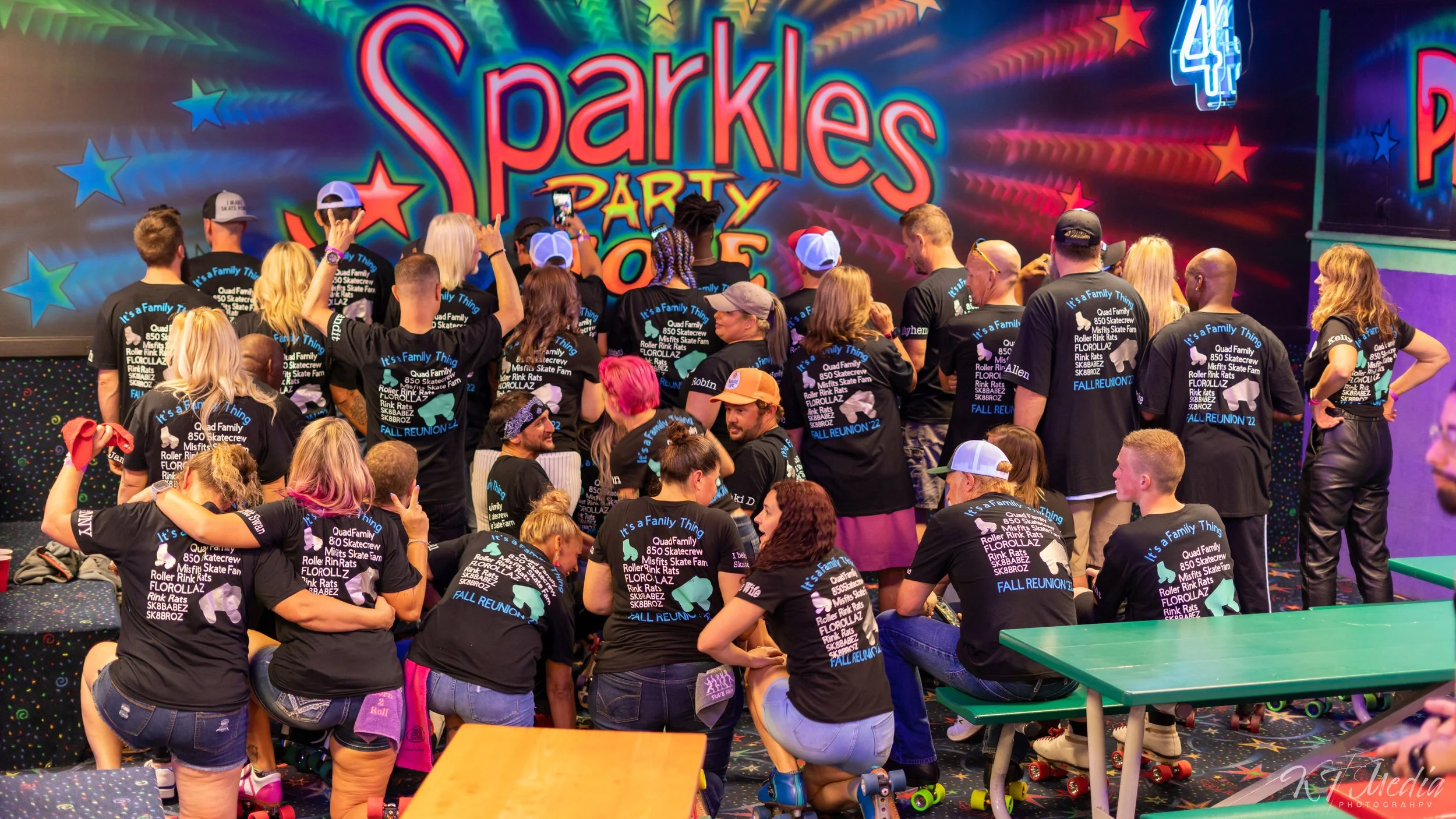 A group of people wearing black T-shirts gathered inside a roller skate rink, with a colorful and vibrant sign that says 'Sparkles Party' in the background.