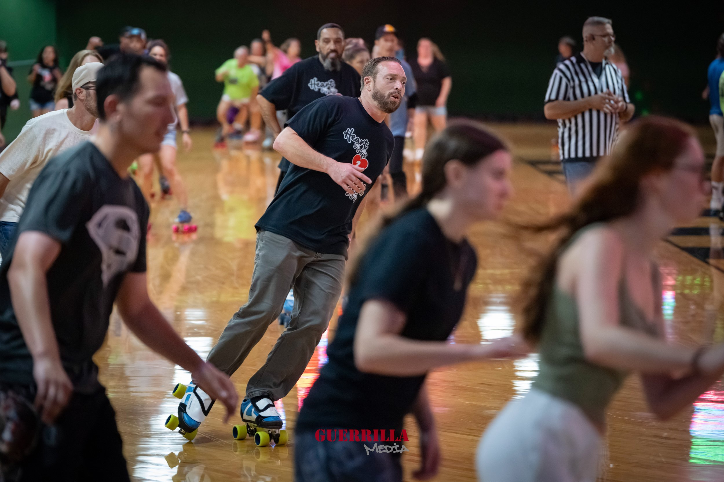 People roller skating indoors, some wearing casual clothing, with a man in focus wearing a black t-shirt and gray pants.