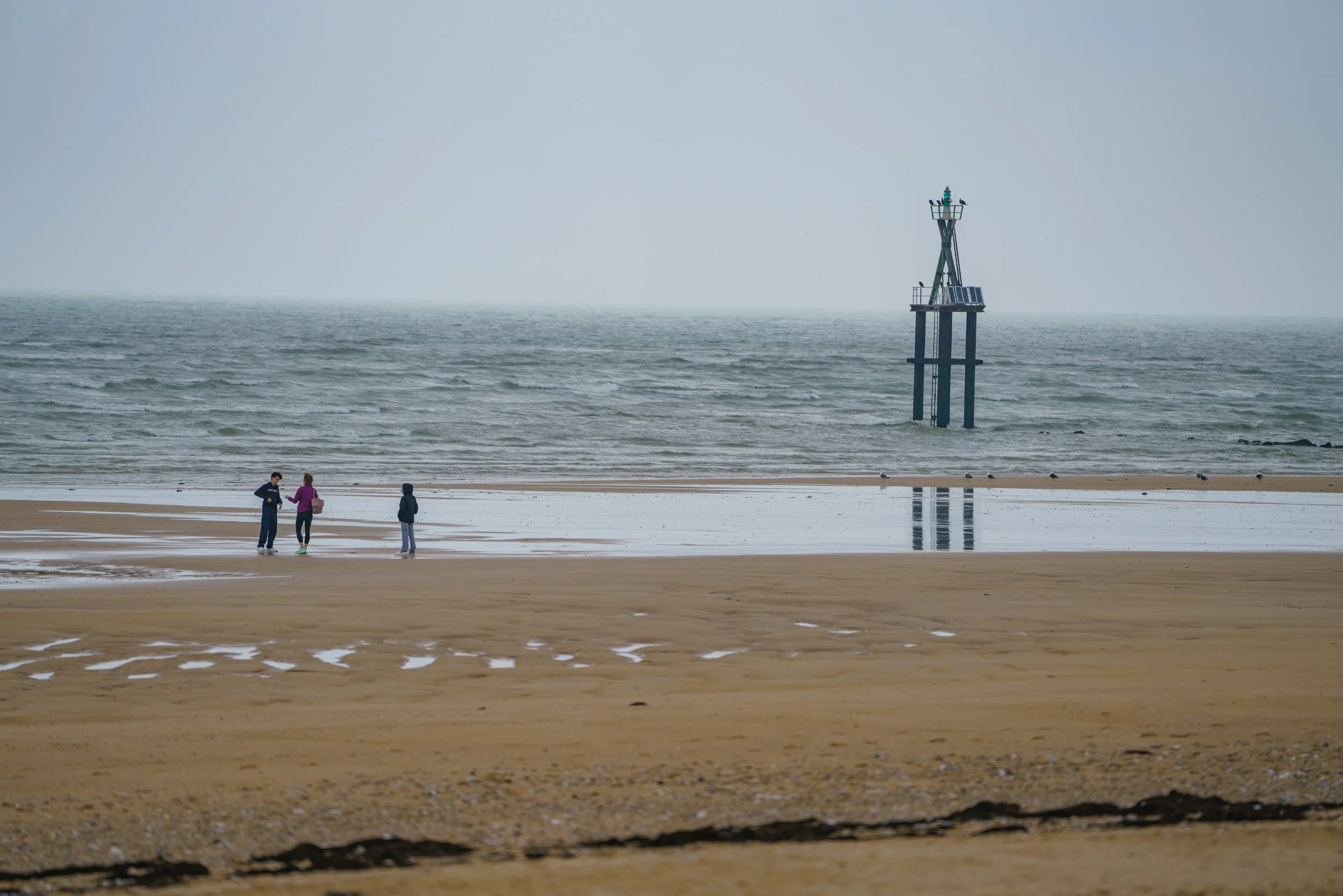 Three people walking on a beach with a lighthouse in the water in the background on a cloudy day.