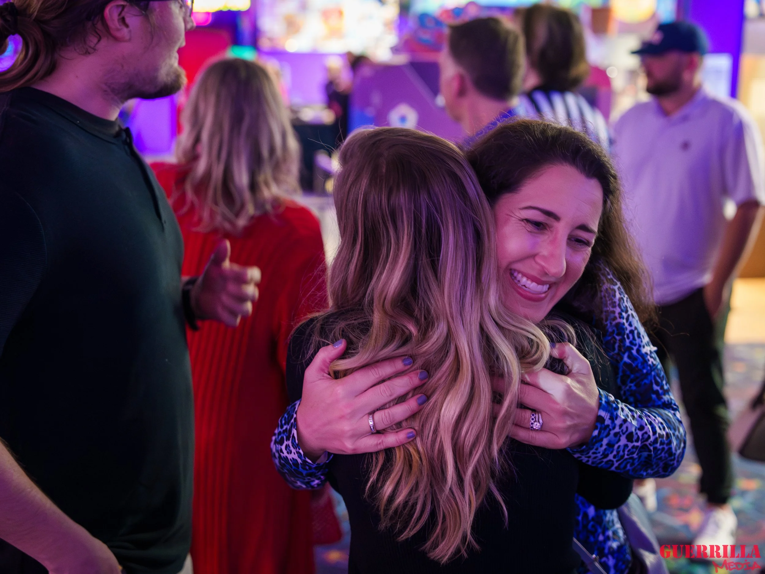 Two women hugging and smiling in a lively, colorful bar or club. Several people are in the background, some talking and others observing. Atmosphere is fun and cheerful.