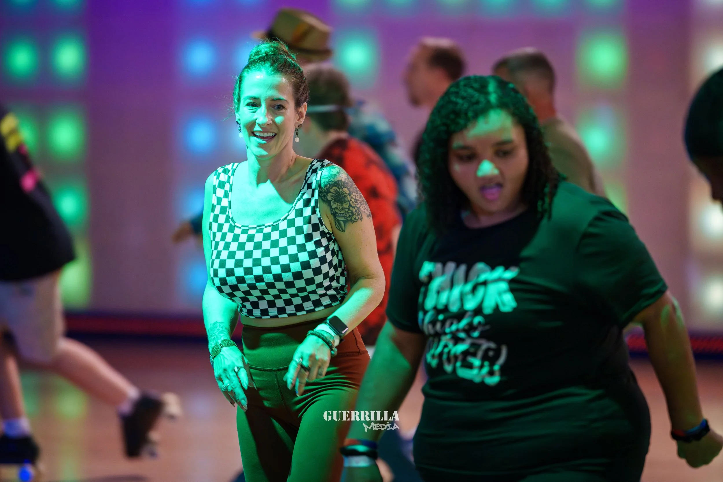Two women dancing at a roller skating rink; one woman in checkered top smiling, the other in a black t-shirt with writing, with colorful lighting in the background.