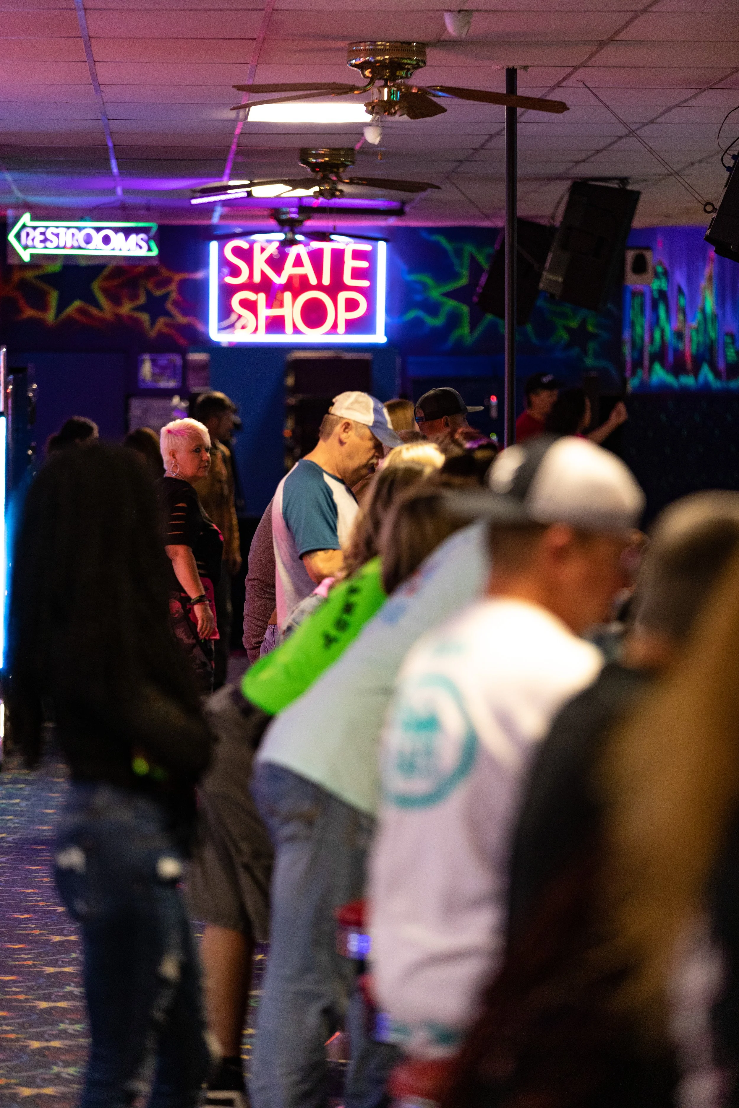 People waiting in line at a roller skating rink with neon signs for restrooms and a skate shop, colorful star mural on the wall, ceiling fans overhead.