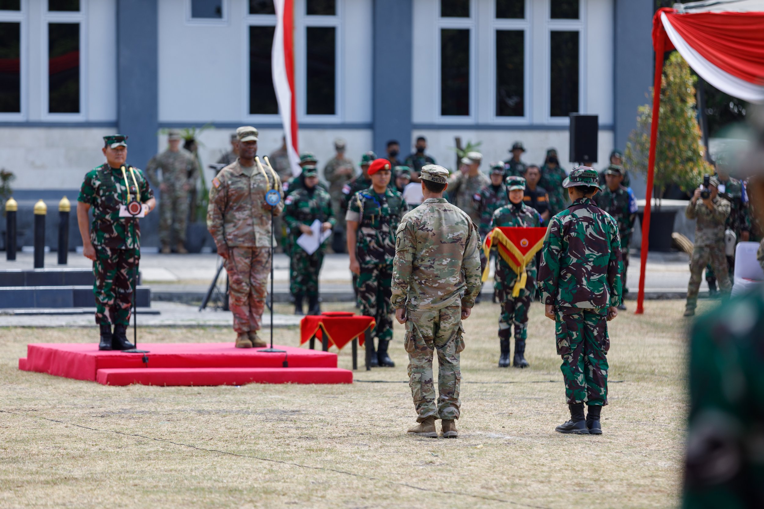 Military personnel in uniform standing at attention during a ceremony on a grassy field, with a building in the background and a red and gold decorated stand to the side.