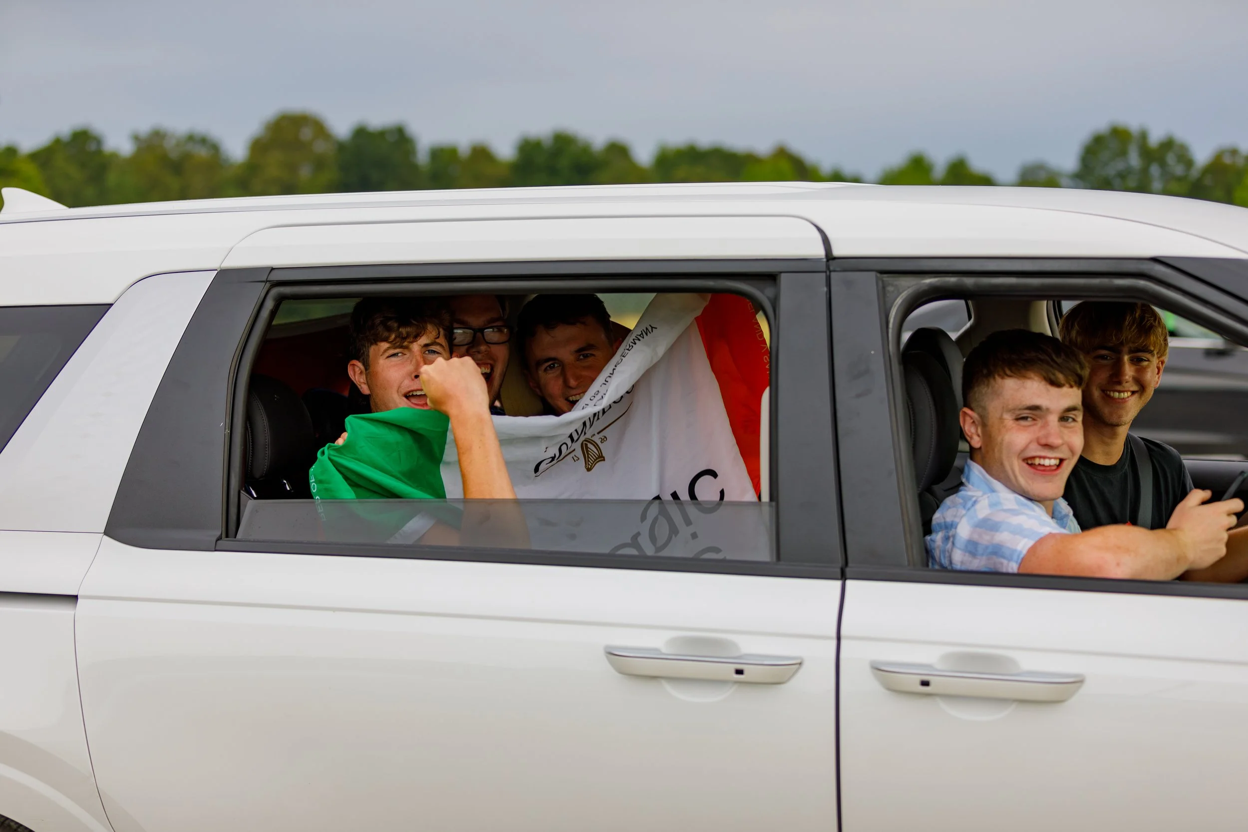 Six young men sitting inside a white vehicle, smiling and celebrating, one of them holding an Italian flag.