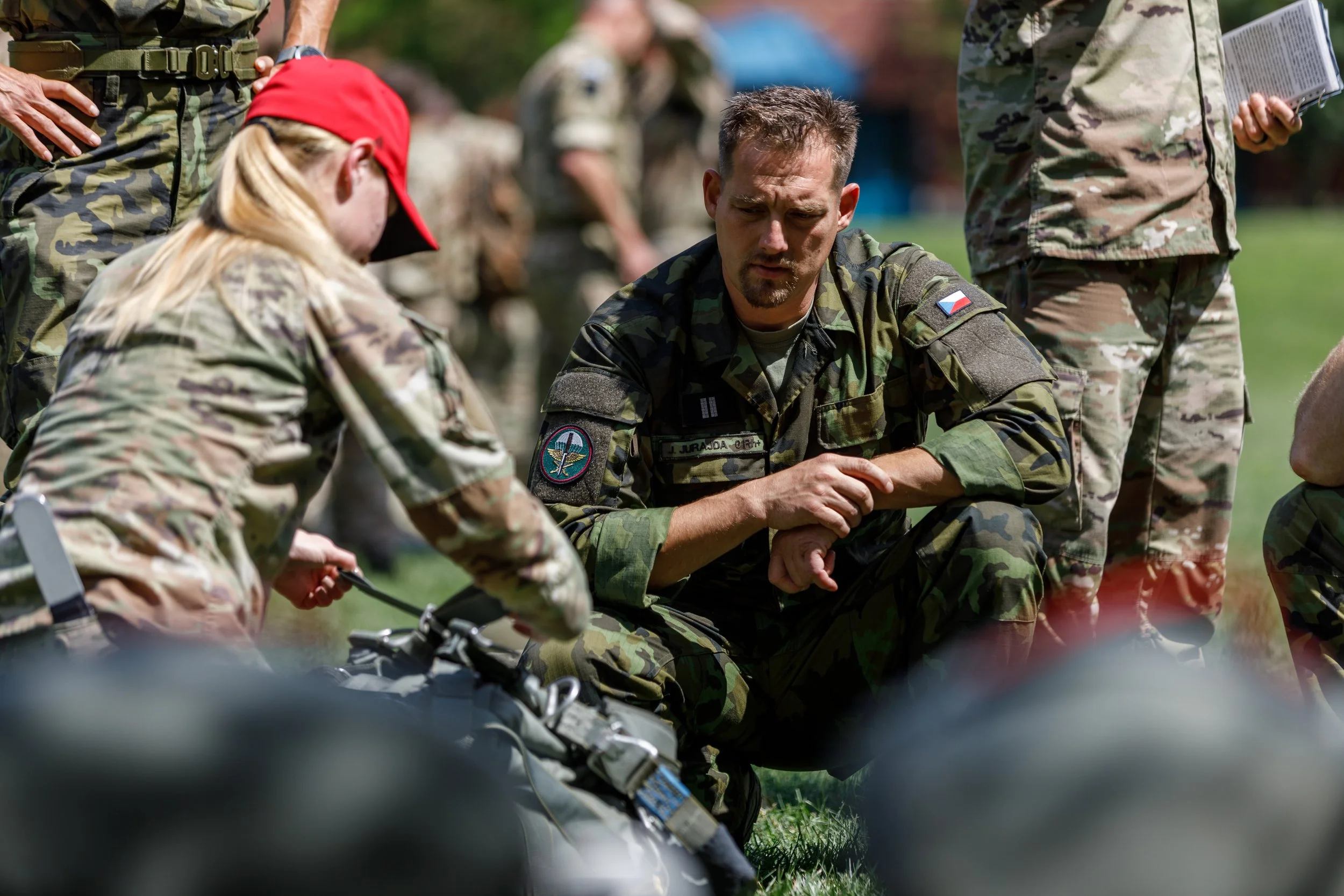 Military personnel, including a man with a Czech flag patch on his uniform, kneeling on the grass and talking to a woman with a red cap and camouflage uniform, with other soldiers in the background.