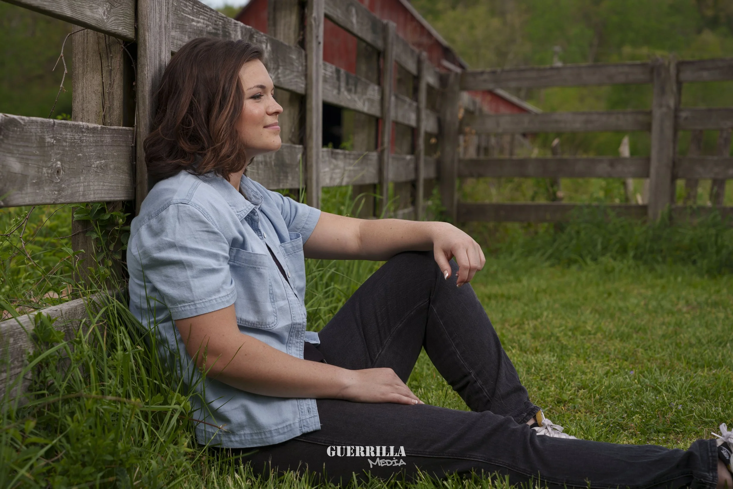 Young woman with brown hair, sitting on grass and leaning against a wooden fence, outdoors on a farm or rural area with green trees and grass in the background.